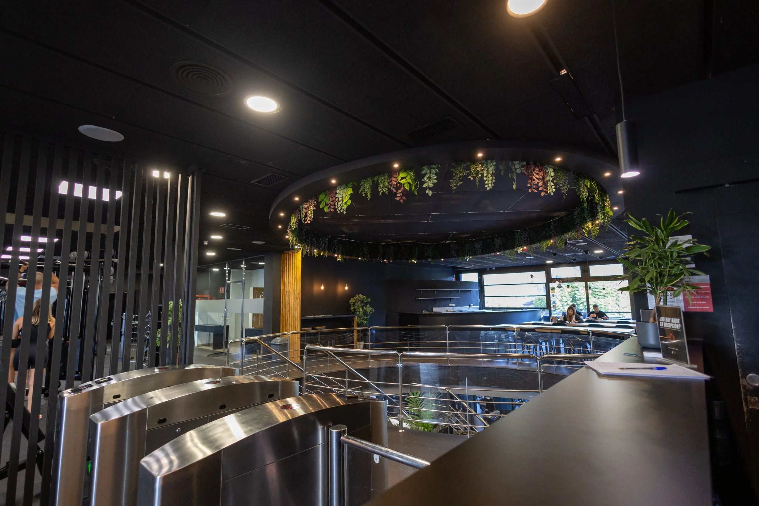 Interior moderno de un espacio público con área de recepción, plantas decorativas y una zona con mesas y sillas junto a ventanas grandes