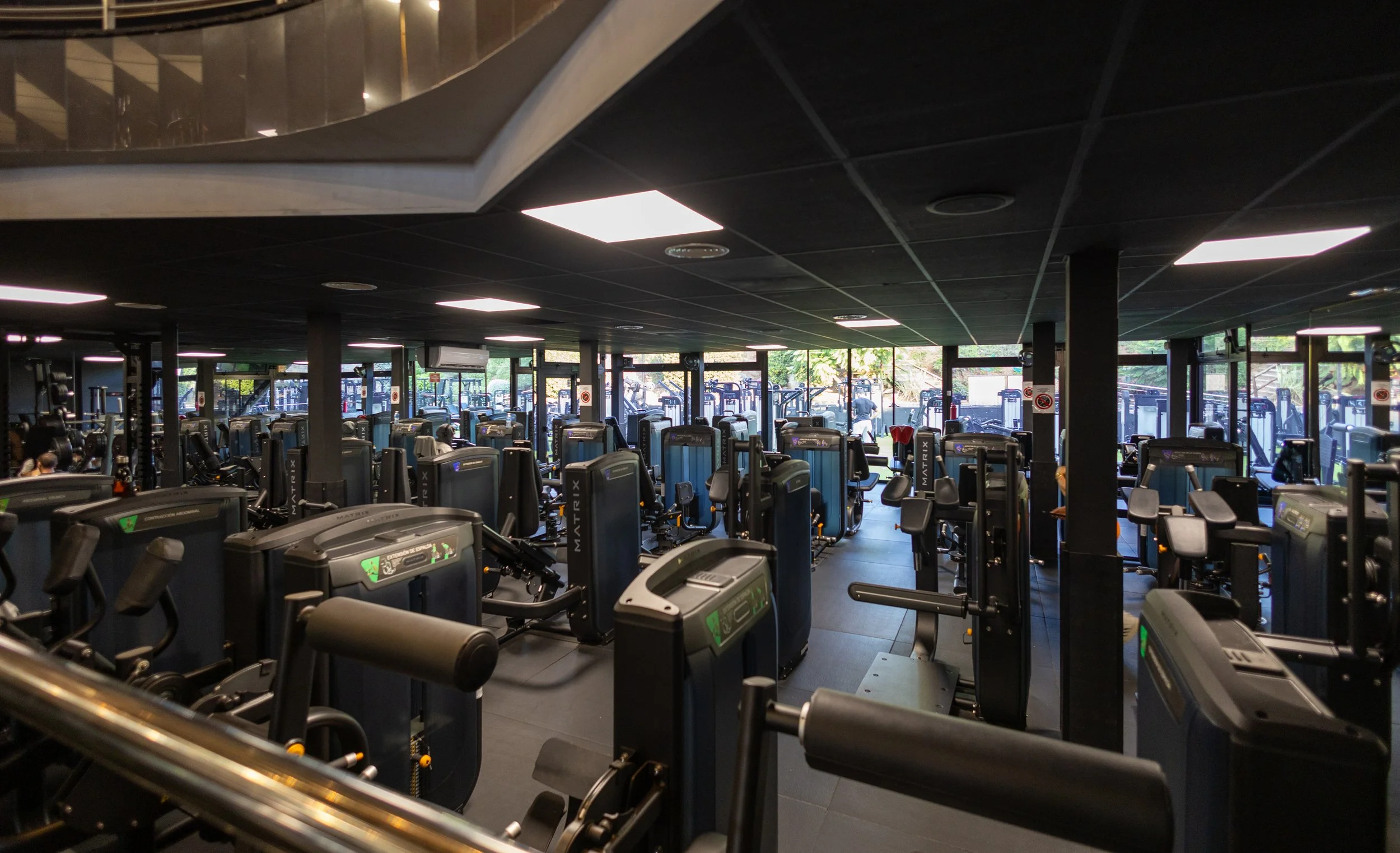 Interior de un gimnasio con varias máquinas de ejercicio y ventanas que dejan entrar luz natural.