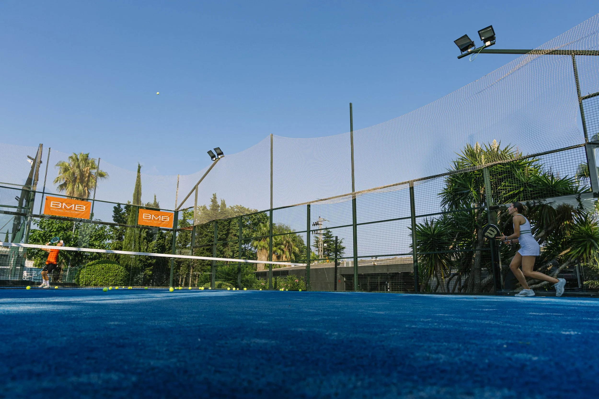 Una cancha de tenis de color azul con dos jugadoras preparadas para jugar, una de ellas en posición de recibir el servicio, en un día soleado con cielo despejado y árboles verdes en el fondo.