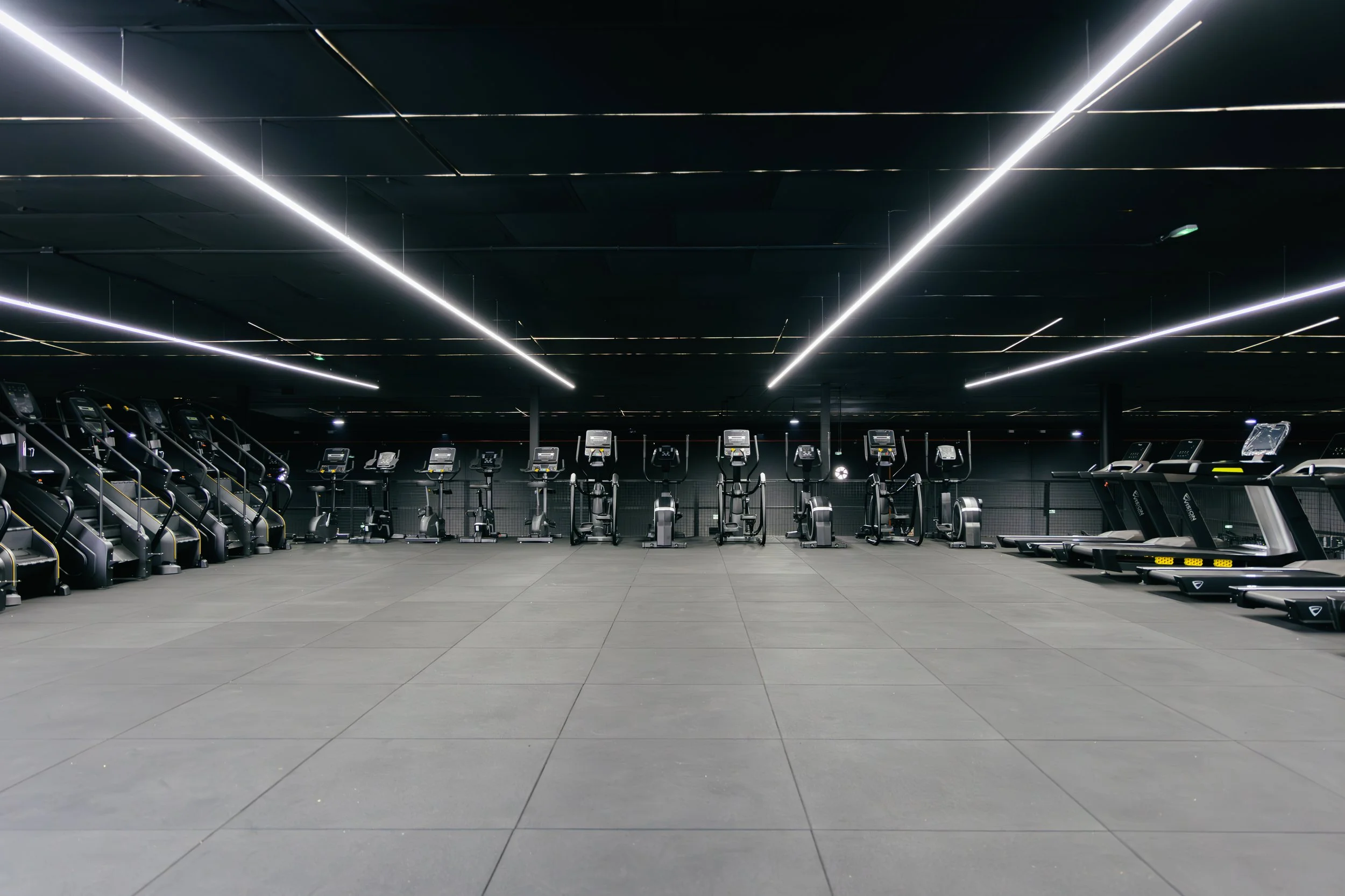 Interior de gimnasio con máquinas de cardio en fila y piso de baldosas grises, iluminación moderna en el techo negro