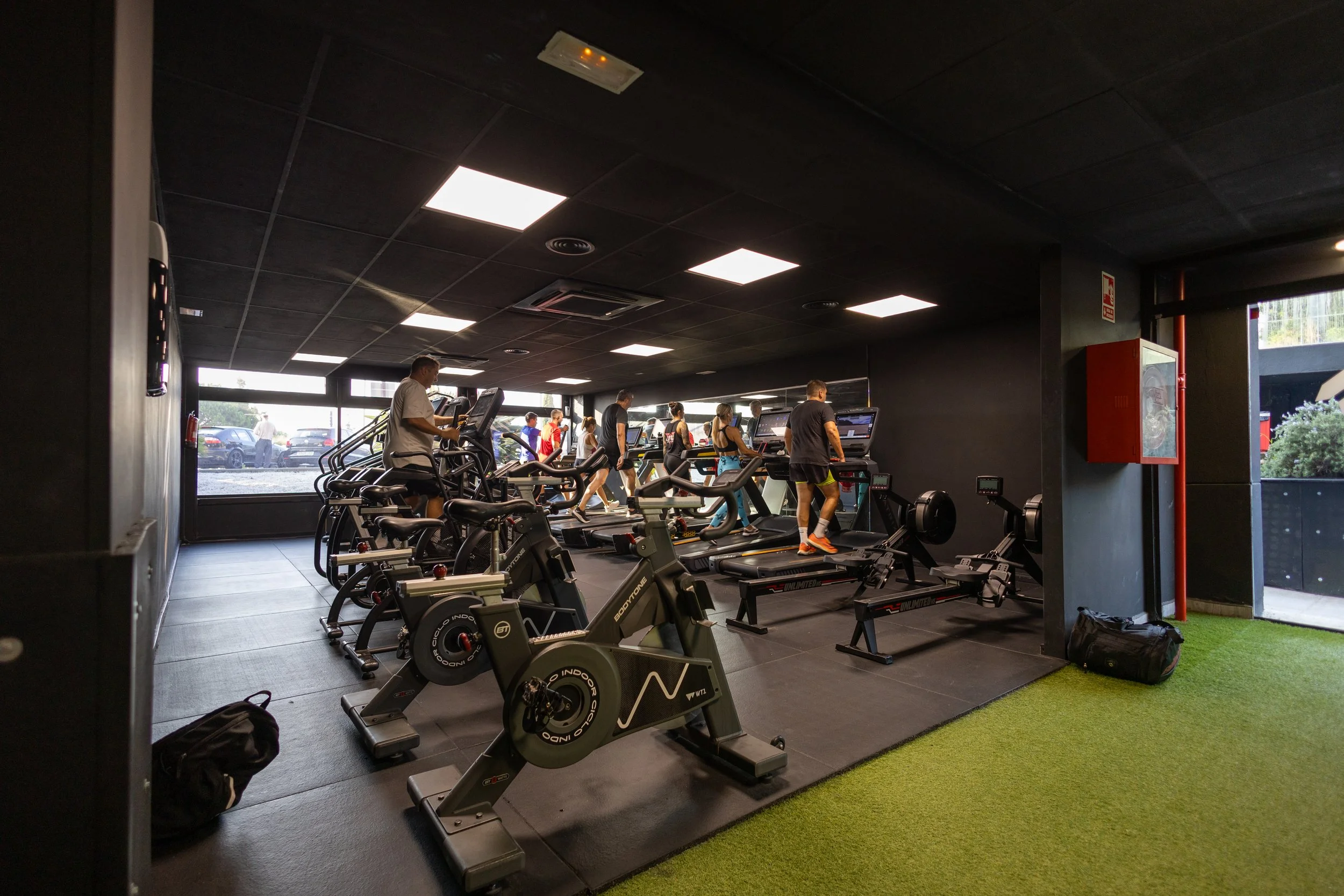 Gimnasio con varias personas haciendo ejercicio en cintas de correr, bicicletas y máquinas de entrenamiento.