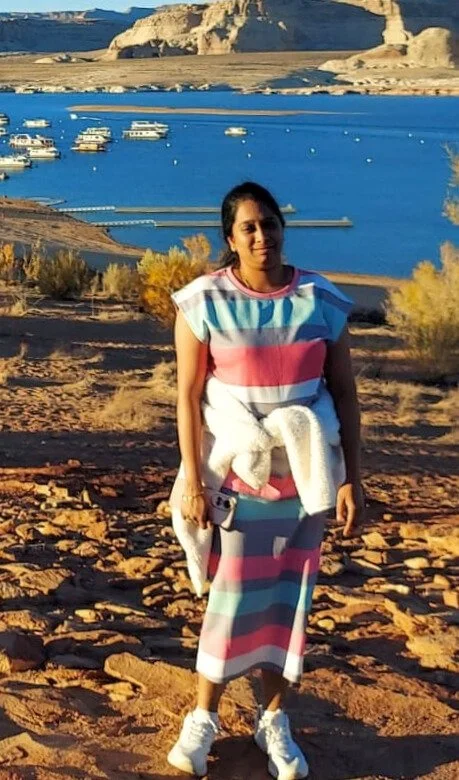 Woman standing on a rocky, sandy terrain near a body of water with boats and a rocky landscape in the background during sunset.