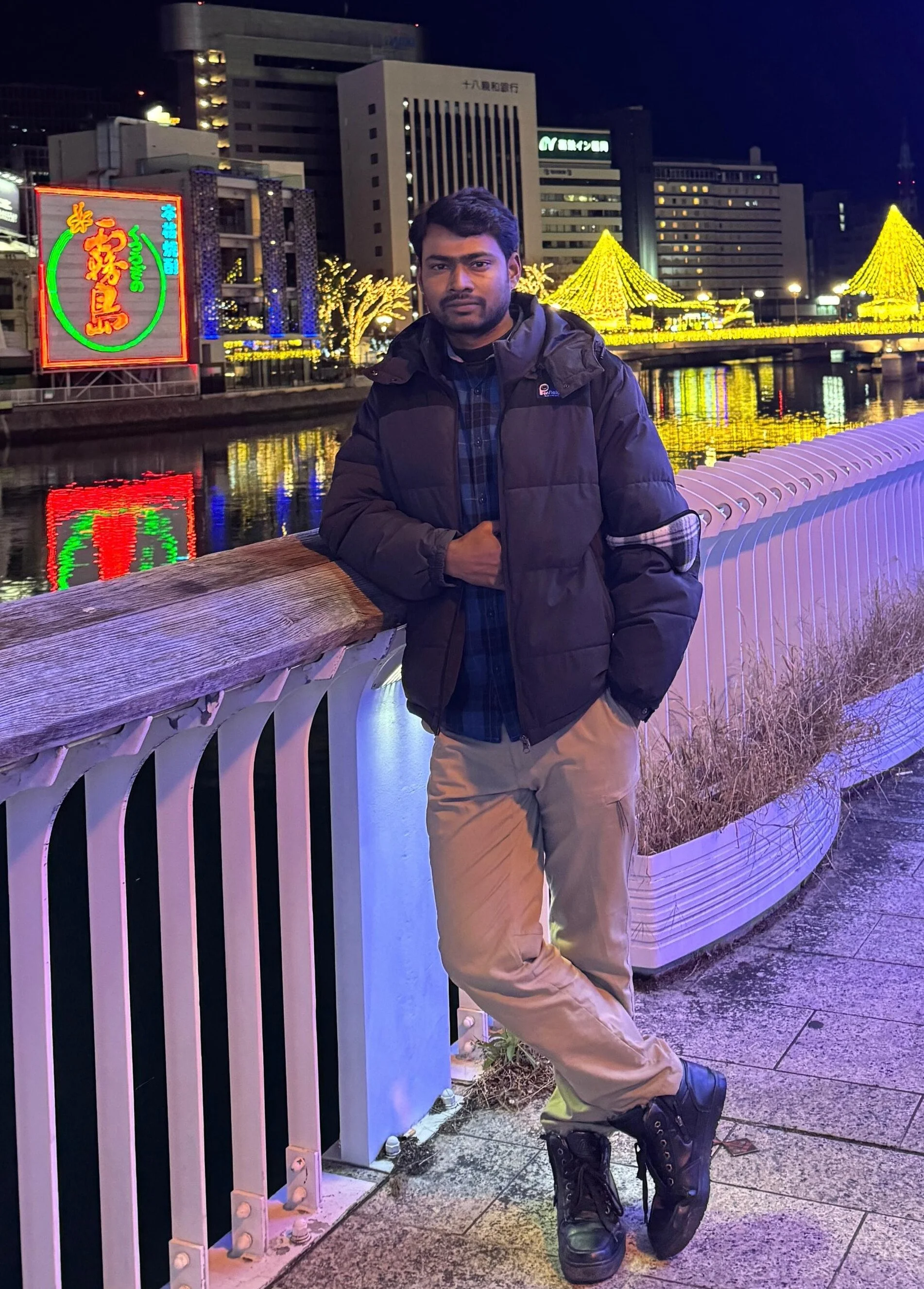 A man standing on a city bridge at night, with illuminated buildings and decorative yellow lights in the background, wearing a dark jacket, plaid shirt, tan pants, and black boots.