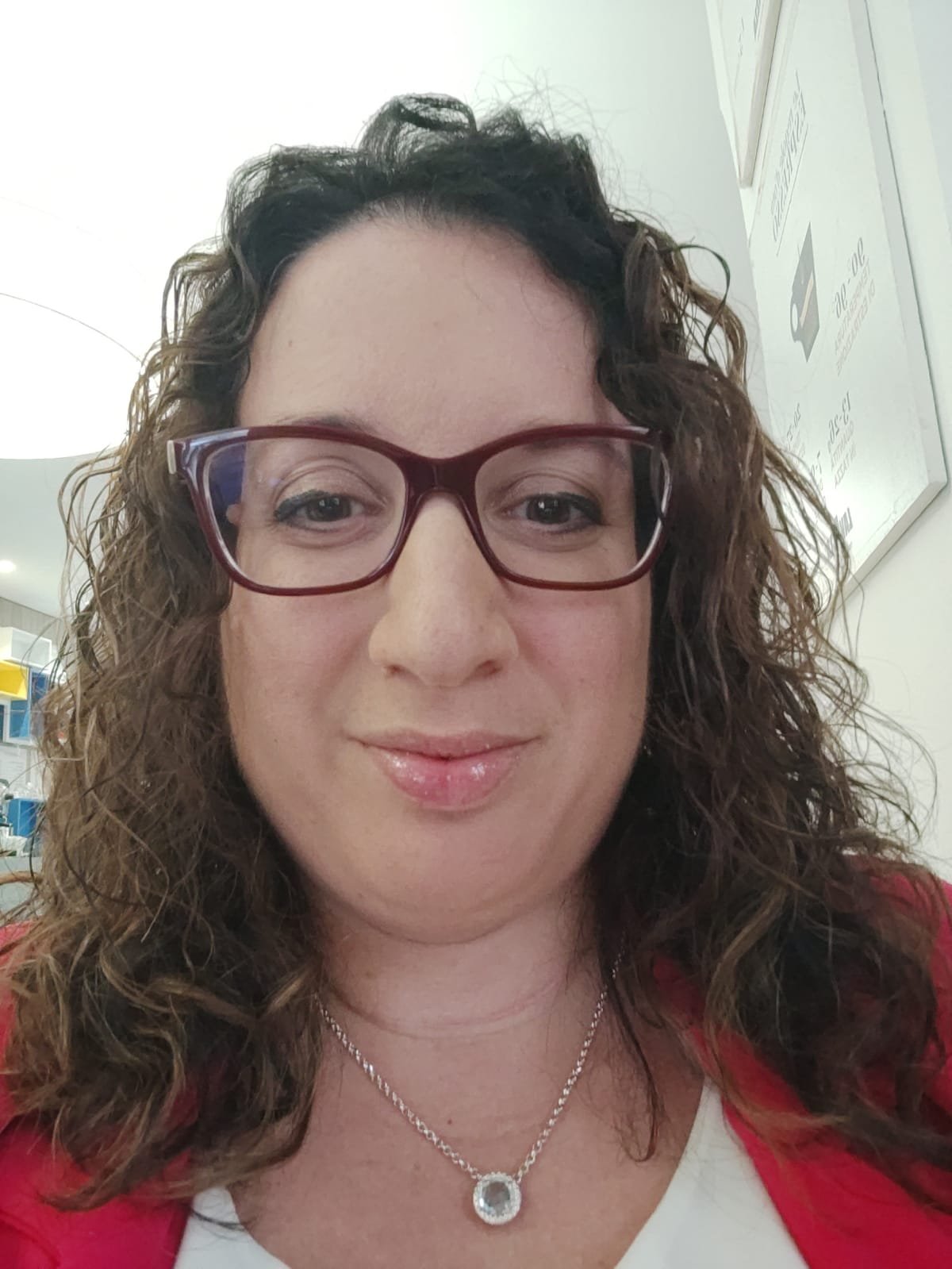 Close-up of a woman with curly hair wearing glasses and a red top, smiling indoors.