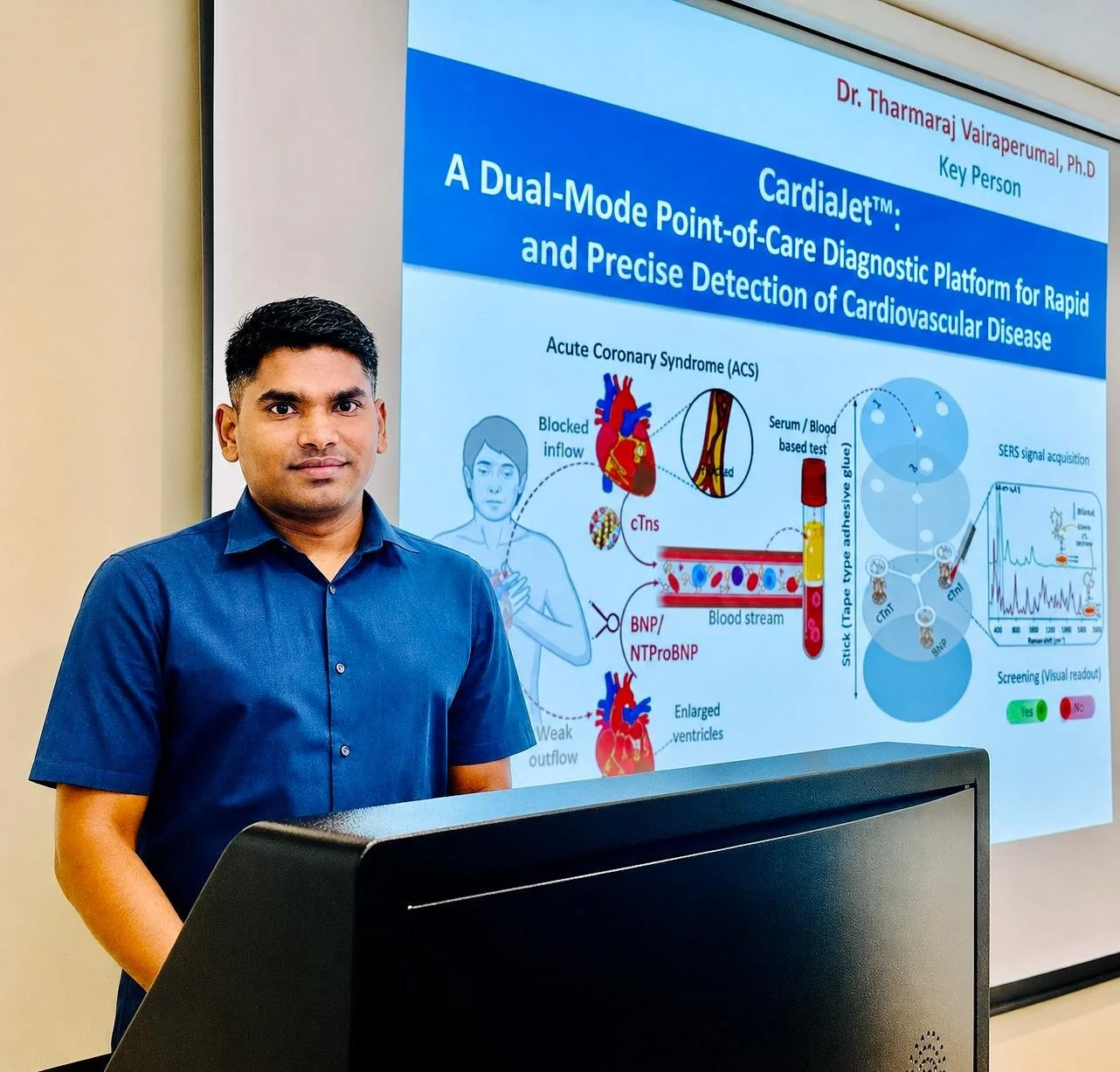 A man in a blue shirt standing in front of a presentation screen displaying a scientific poster on cardiovascular disease detection using CardioJet technology.