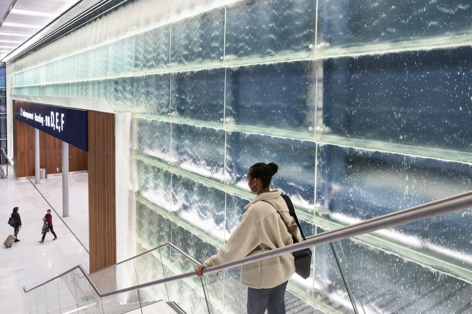 Une femme portant un manteau beige, des créoles, et un sac à l'épaule, se tient sur un escalier dans un aéroport, regardant vers le bas, avec un mur en verre en arrière-plan.