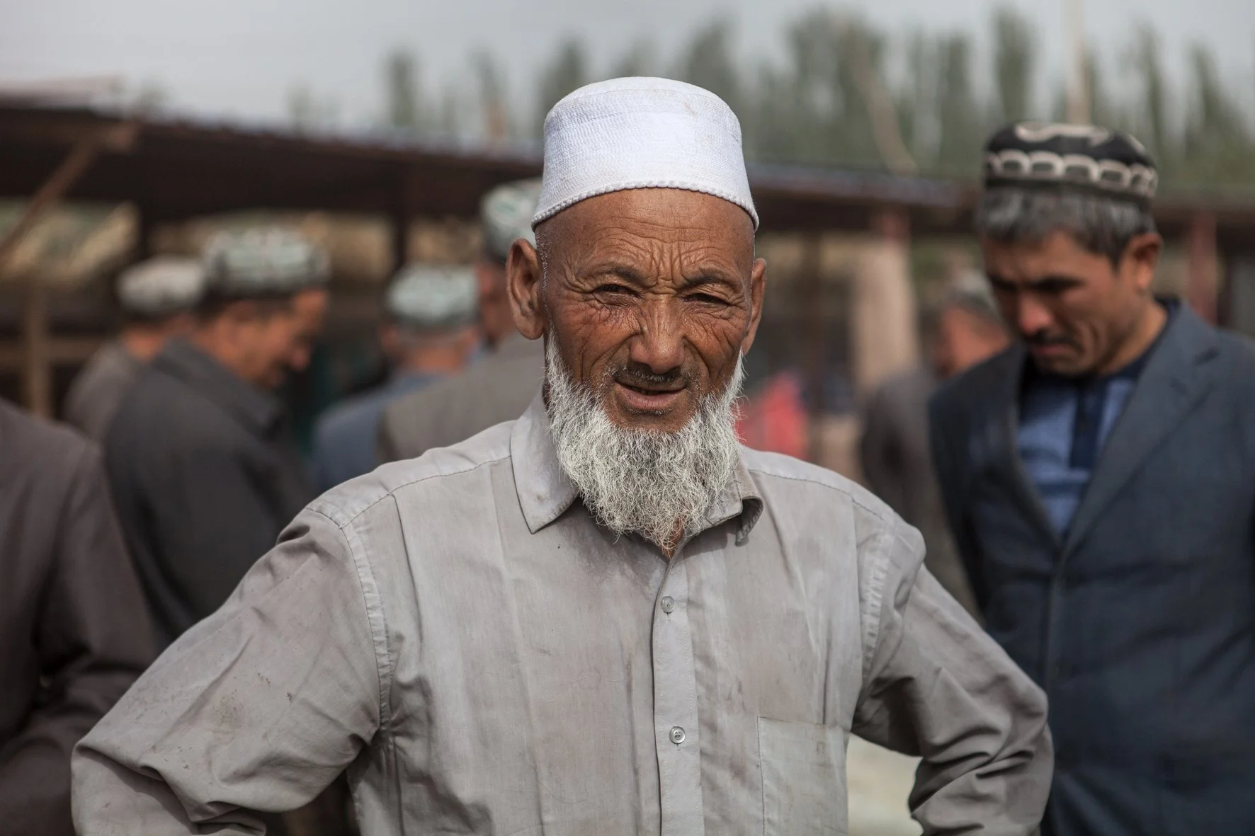 Un homme âgé avec une barbe blanche et une coiffe blanche regarde l'objectif, avec d'autres personnes en arrière-plan à Kashgar en Chine.
