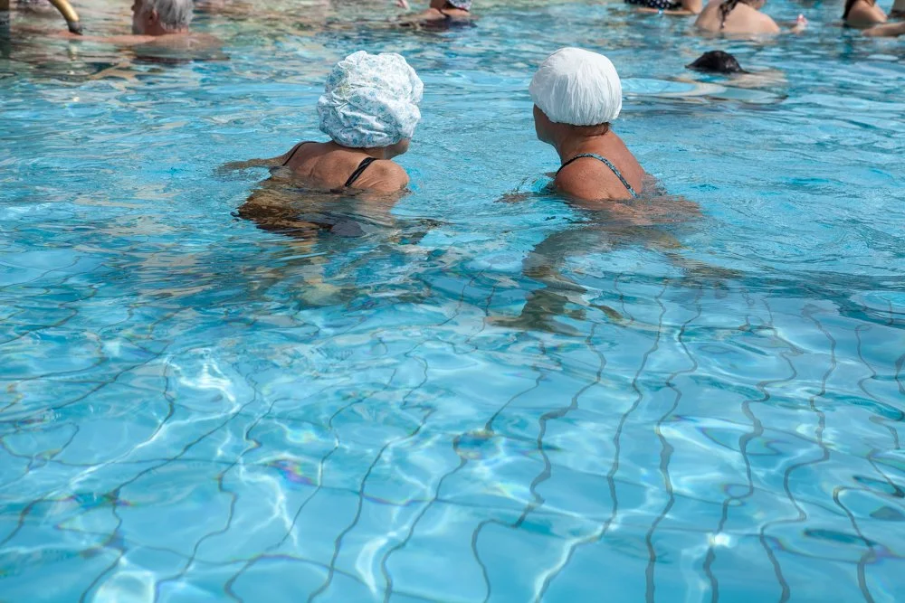 Deux personnes âgées portant des bonnet de bain, nageant ou discutant dans une piscine.