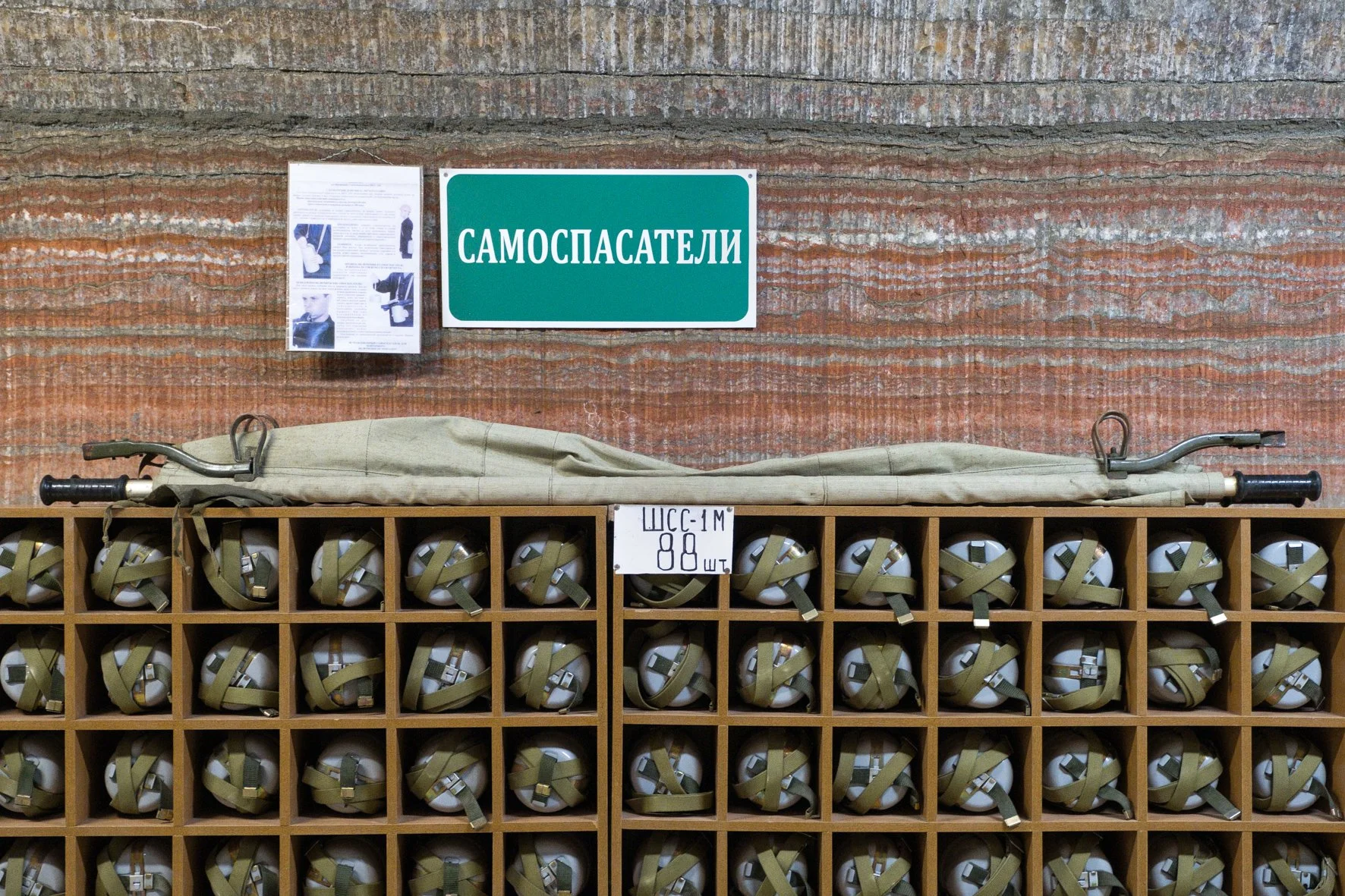 Une étagère contenant de nombreux masques respiratoires avec des sangles vertes, un lot de 88, un panneau en ukrainien avec le mot "самоспасатели" et des informations en russe, un mur de roche rouge en arrière-plan.