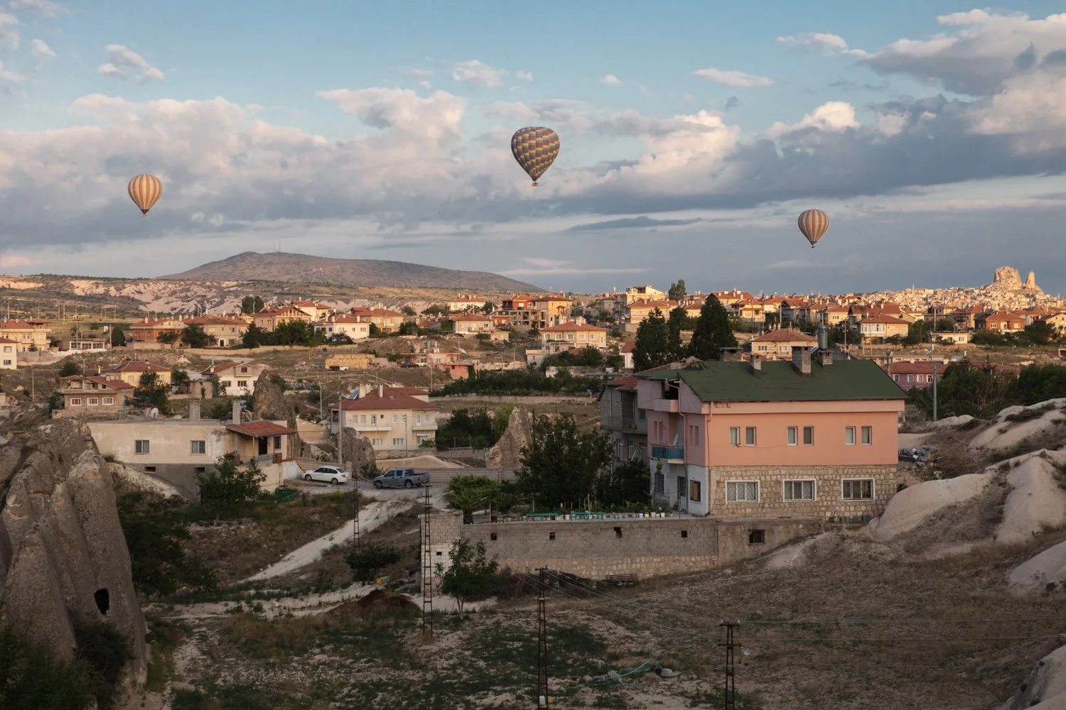 Vue panoramique d'un paysage urbain avec plusieurs maisons et bâtiments, plusieurs montgolfières dans le ciel, montagnes en arrière-plan, ciel nuageux avec un peu de lumière