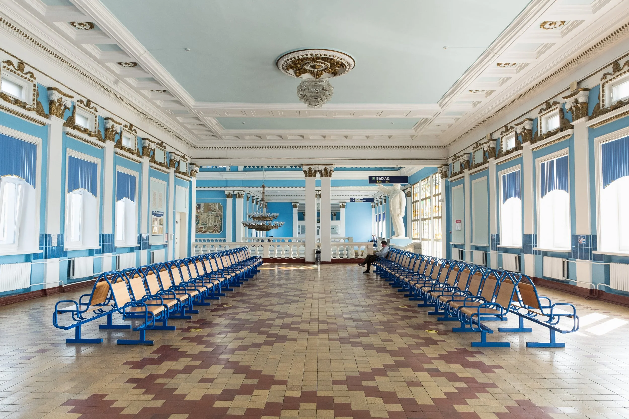 Un hall d'attente spacieux avec des fenêtres, des chaises bleues en rangée, un lustre en cristal suspendu, une statue blanche de Lénine dans un coin, et des panneaux de sortie en russe dans une gare Biélorusse.