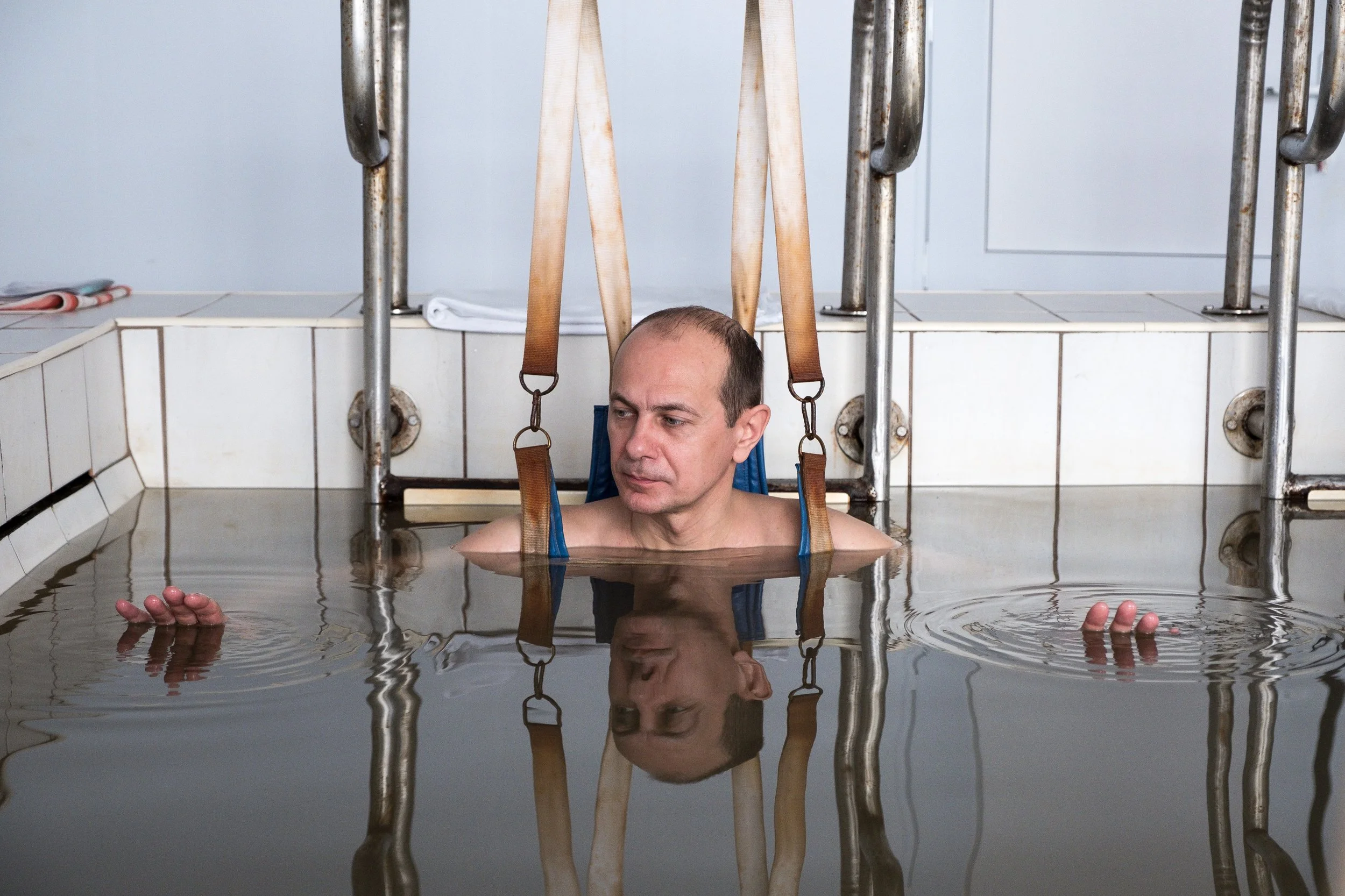 Un homme dans une piscine d'eau sombre, regardant sur le côté, avec des sangles attachées à une structure métallique le maintenant au-dessus de lui.