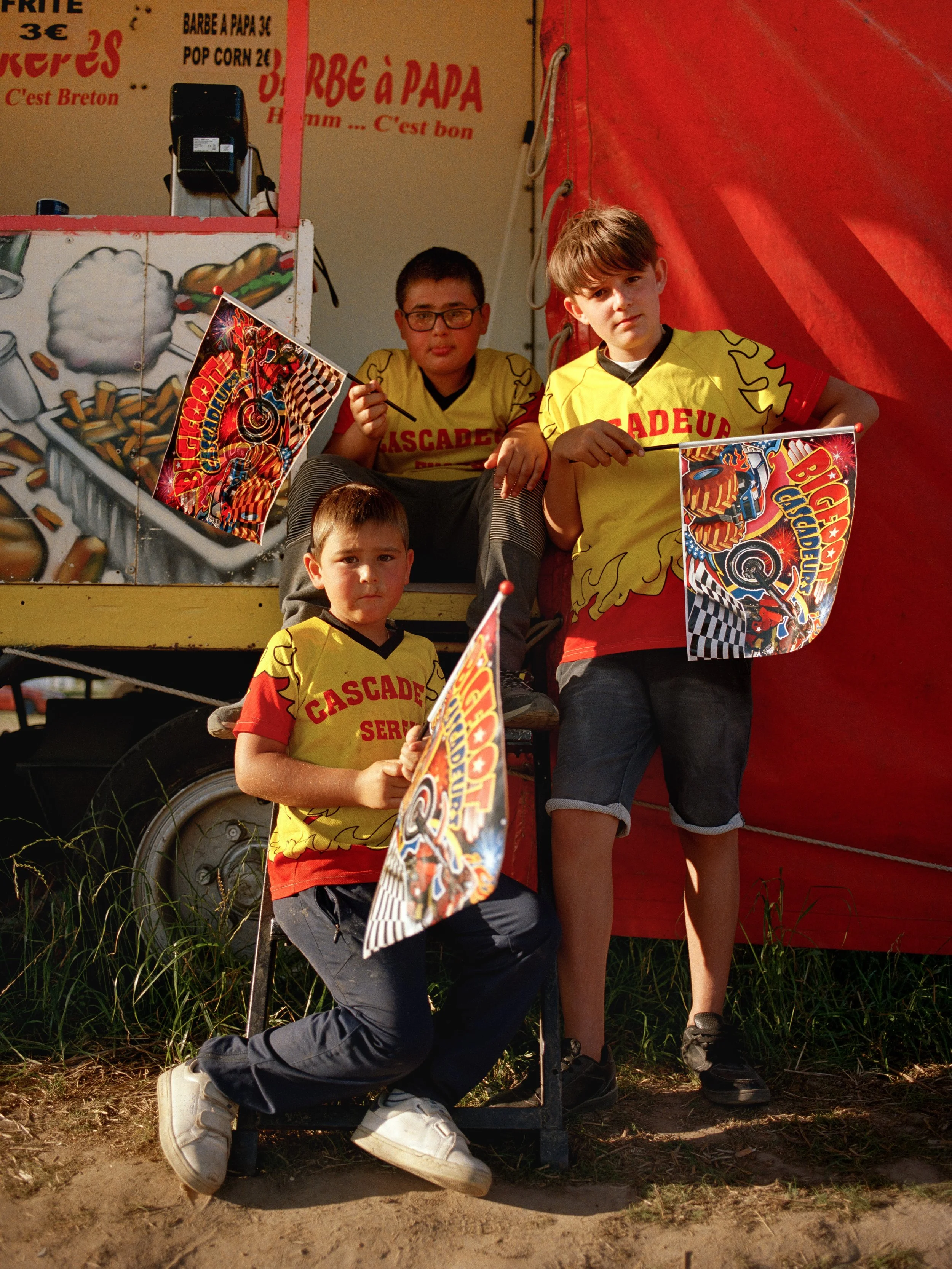 Trois garçons jeunes posant avec des drapeaux devant une roulotte. Deux d'entre eux sont assis, le dernier est debout, tous portant des t-shirts jaunes avec la mention "Cascade".