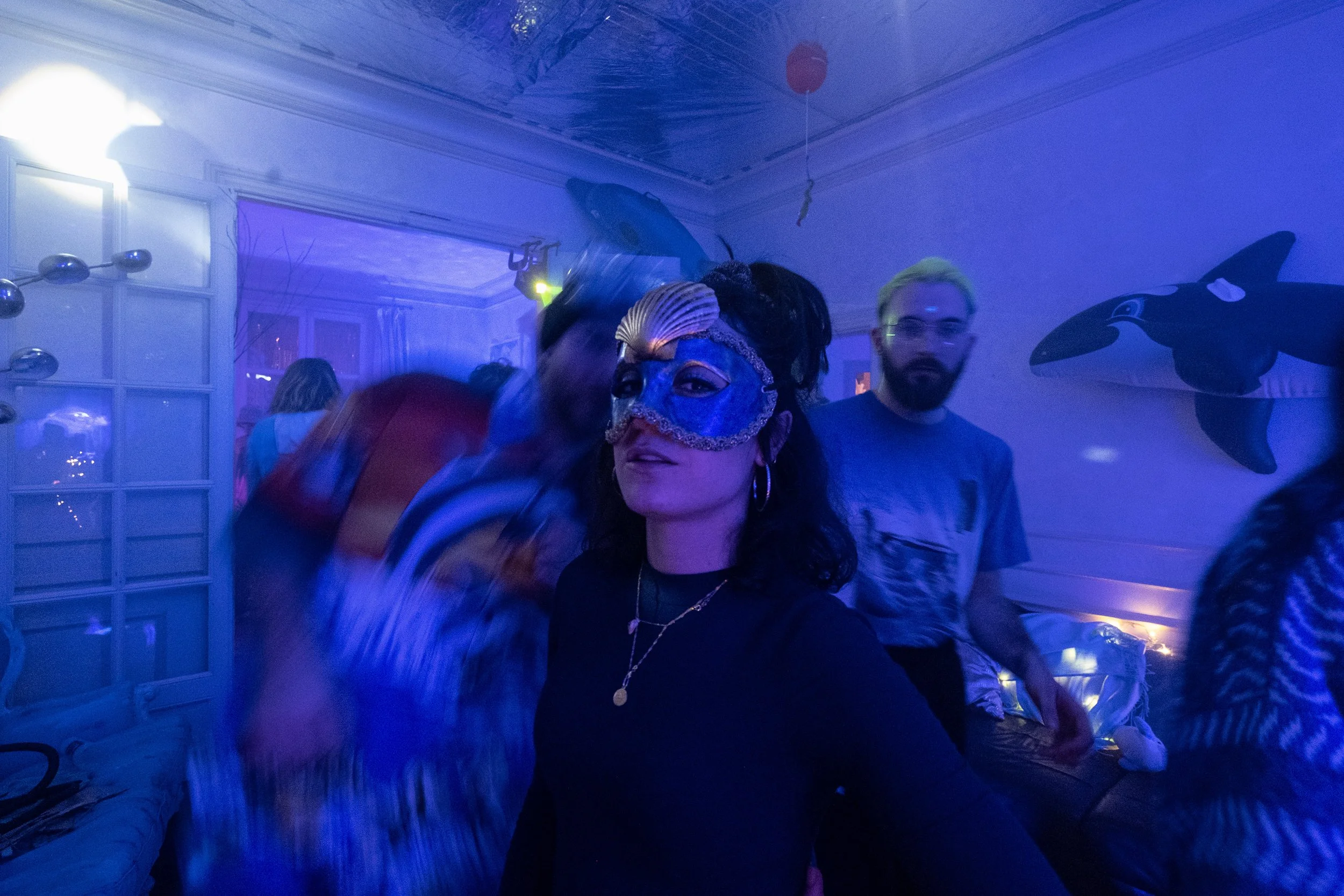 Une femme porte un masque bleue décoré de coquillage lors d'une fête avec ambiance bleutée, entourée de plusieurs personnes dans une pièce décorée avec un orque gonflable et un poisson rouge volant en décoration.