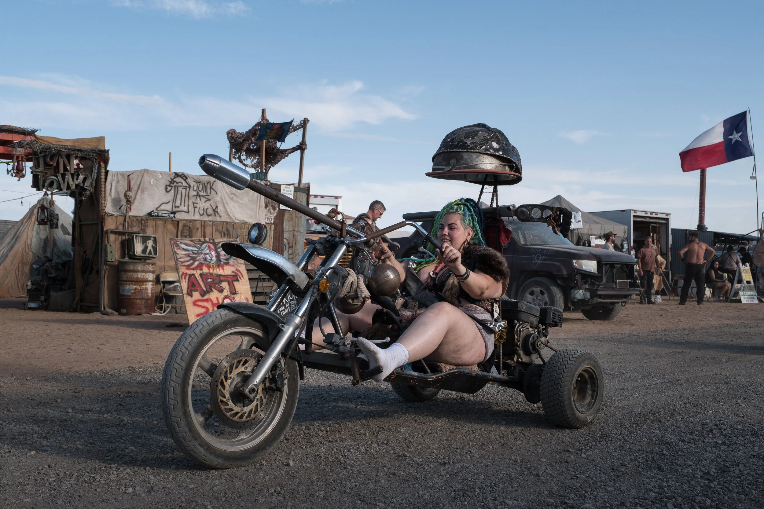 Une femme à cheveux colorés, souriante, assise sur un véhicule modifié ressemblant à une moto avec trois roues, dans un environnement désertique avec des tentes, des véhicules, des personnes et un drapeau du Texas en arrière-plan à Wasteland.