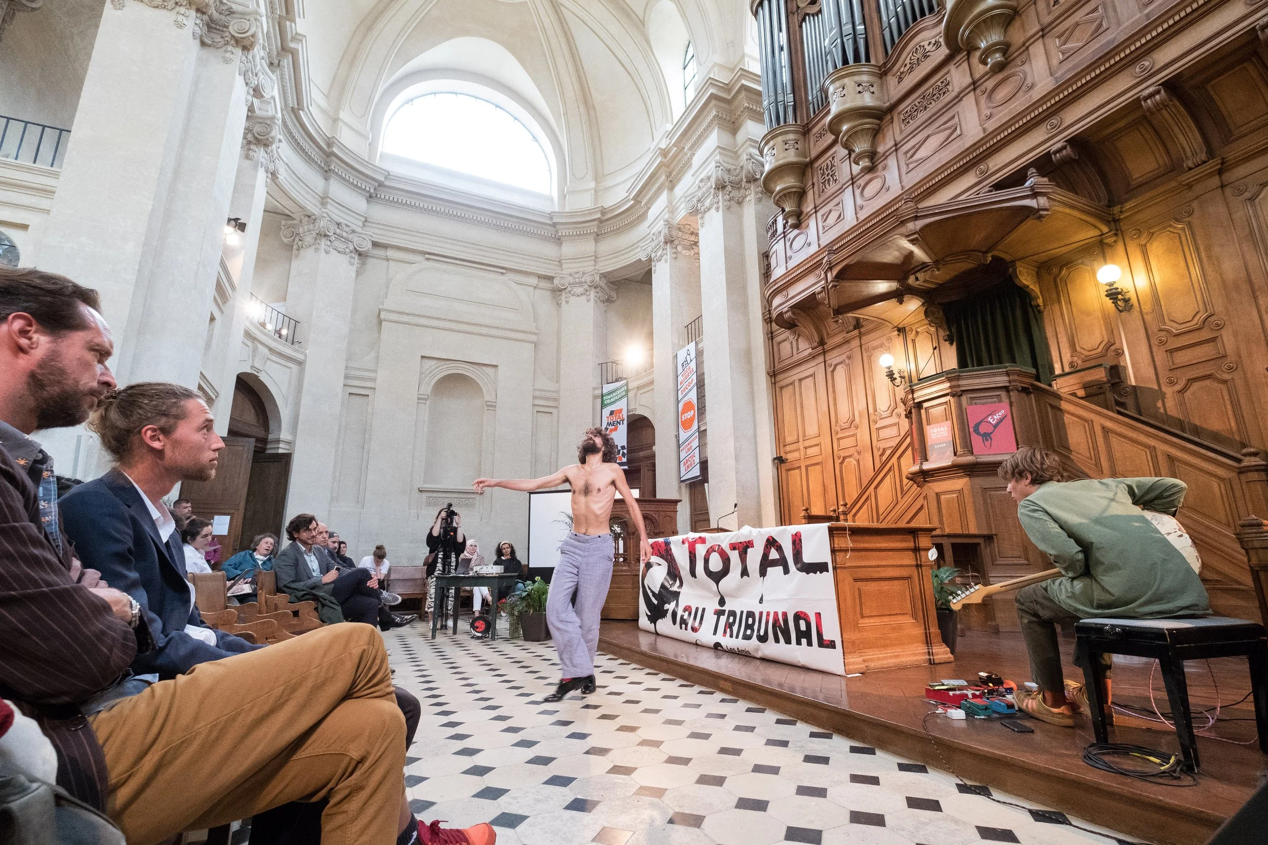 Une action militante de 350.org dans une église, où un homme torse nu danse devant une audience et un musicien joue de la guitare électrique. Un panneau avec le texte 'TOTAL' et 'TRIBUNAL' est présent près de la scène.