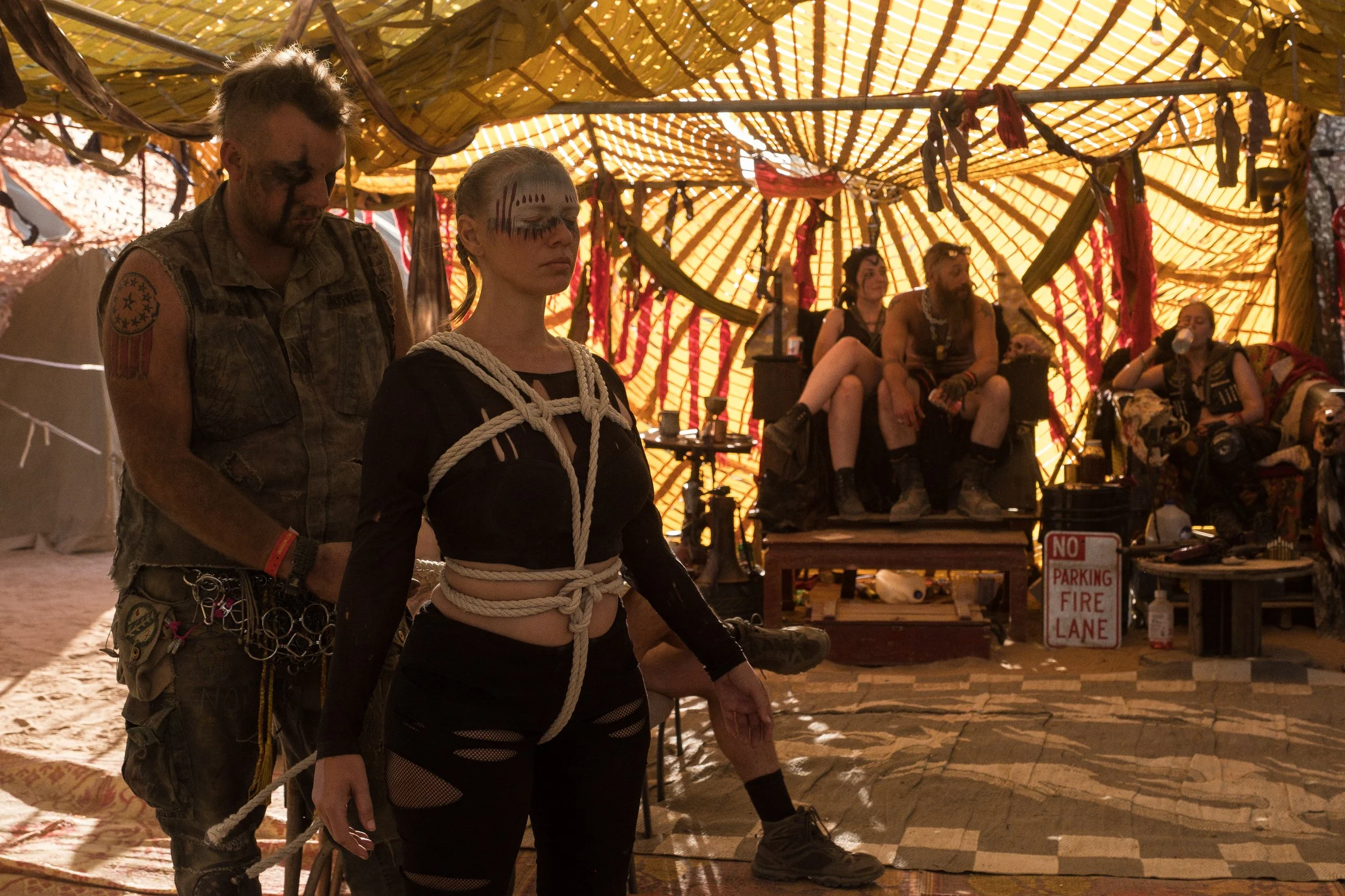 Un groupe de personnes déguisées avec un style post-apocalyptique, certains enchainés ou avec du maquillage sombre, à l'intérieur d'une tente décorée avec des tissus colorés en motif rayé, ambiance sombre et décontractée à Wasteland.