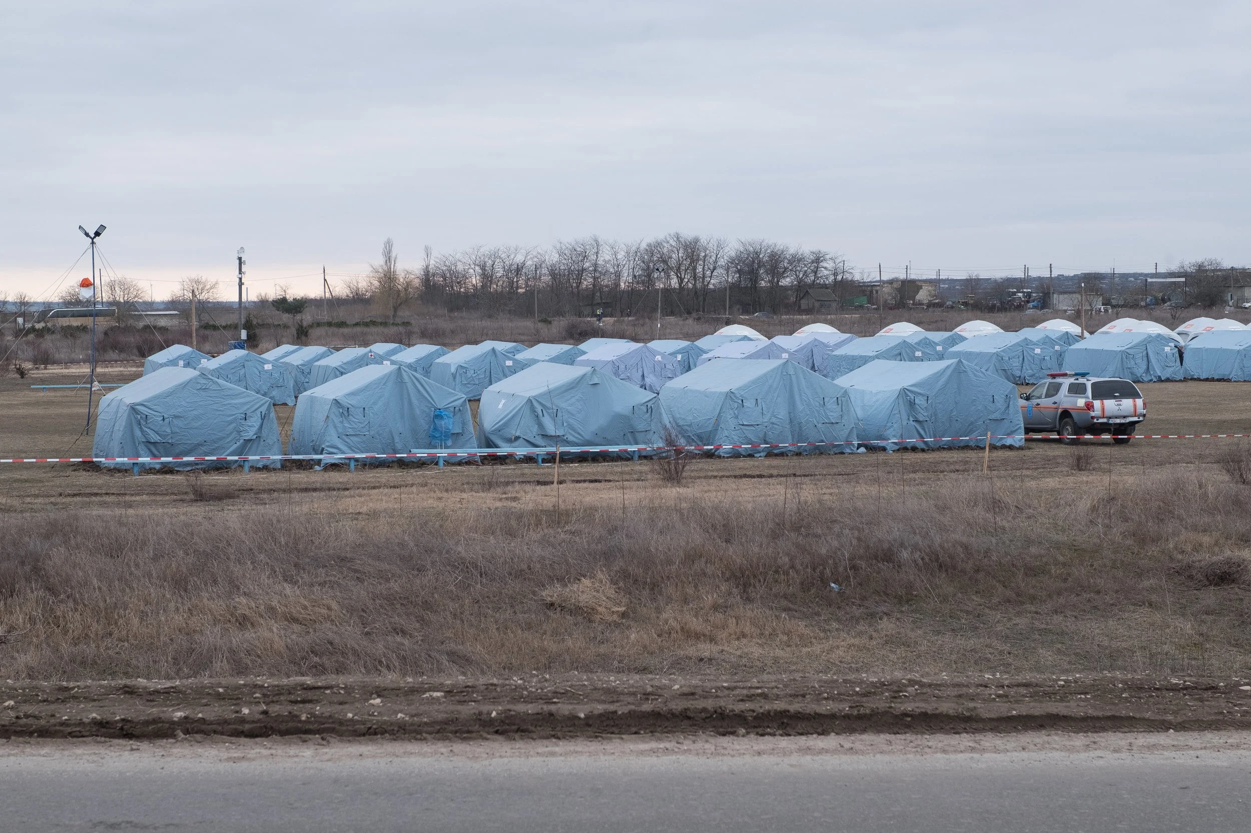 Tentes de secours bleues sur un terrain plat, entourées d'une barrière de signalisation, avec un véhicule de police à droite, dans un paysage rural sous un ciel nuageux.