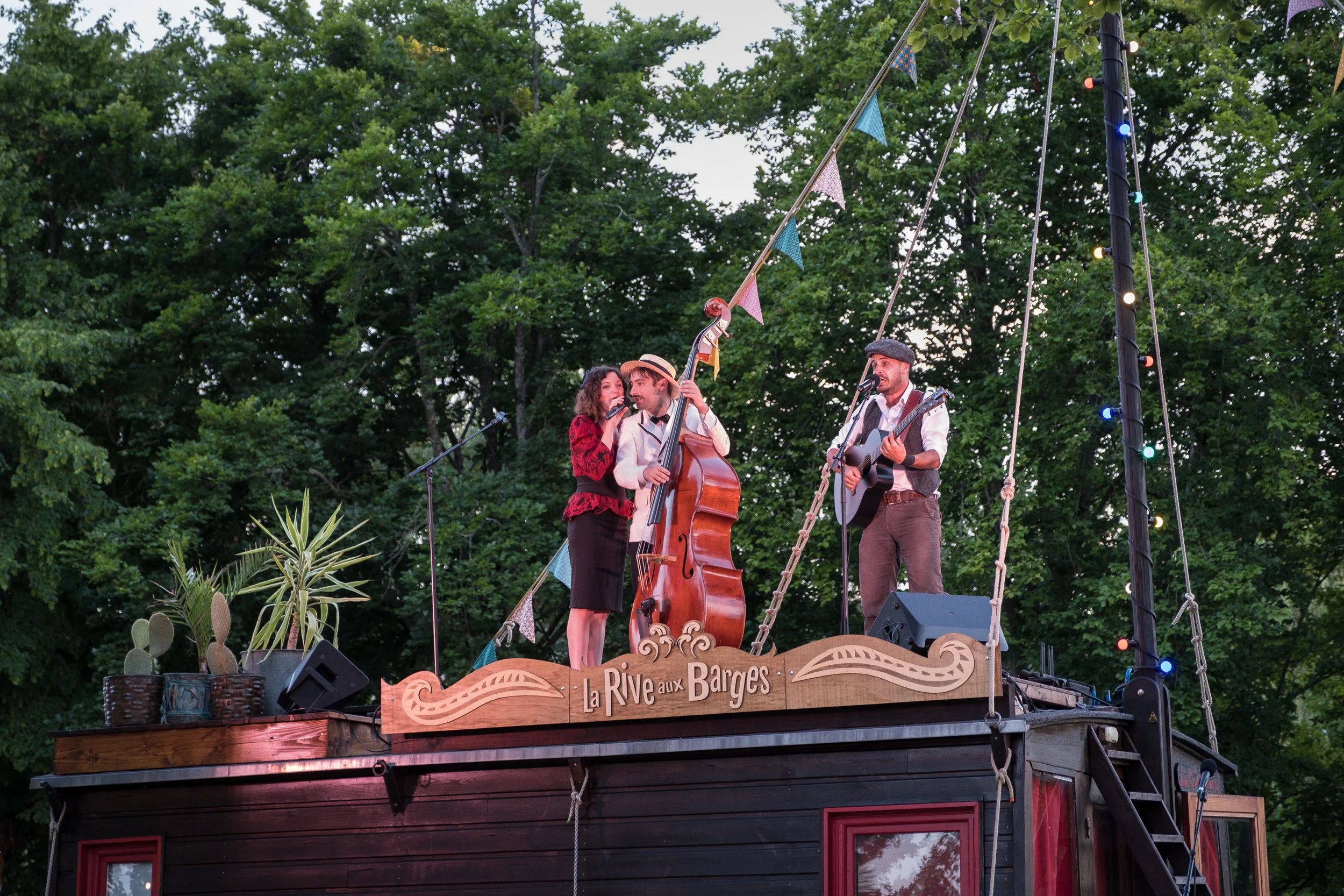 Trois musiciens jouent sur une scène en plein air, avec un fond d'arbres. Un homme joue de la guitare, une femme chante, un autre homme joue du violoncelle. La scène est décorée avec des drapeaux colorés et une pancarte "La Rive aux Barges".