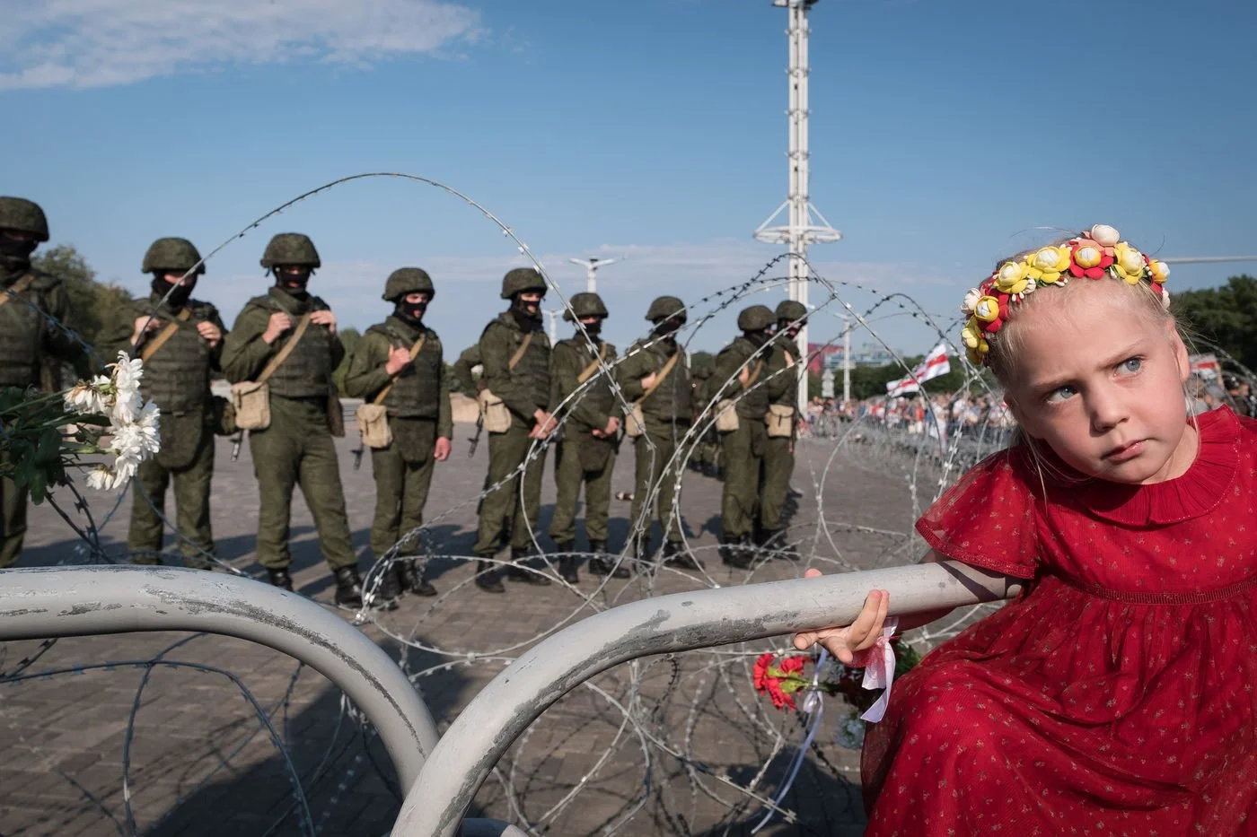 Une petite fille en robe rouge et couronne de fleurs, le regard dans le vague devant une barrière de fil de fer barbelé, avec un groupe de soldats en uniforme en arrière-plan lors d'une manifestation à Minsk, en Biélorussie.