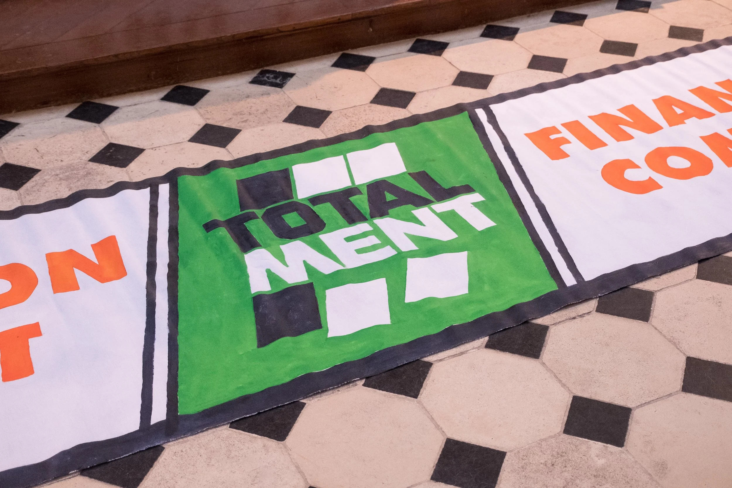 Un panneau peint avec le texte "TOTAL MENT" en vert et noir, entouré d'une bordure noire, posé sur un sol en carreaux blancs et noirs.