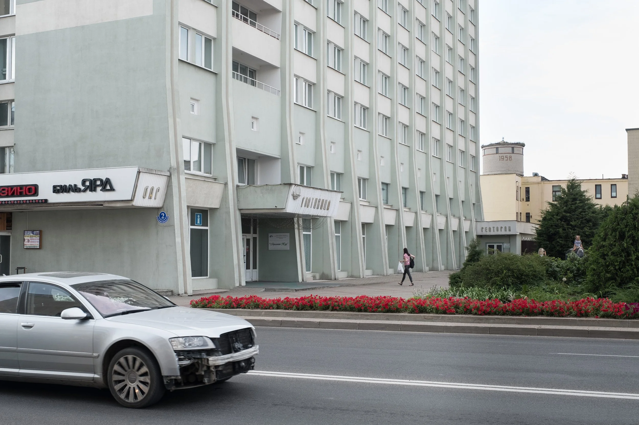 Une rue urbaine en Biélorussie avec un immeuble résidentiel, une voiture accidentée roulant et une femme au loin. L'immeuble est vert clair et a plusieurs fenêtres. Il y a des arbres et des fleurs devant l'immeuble.