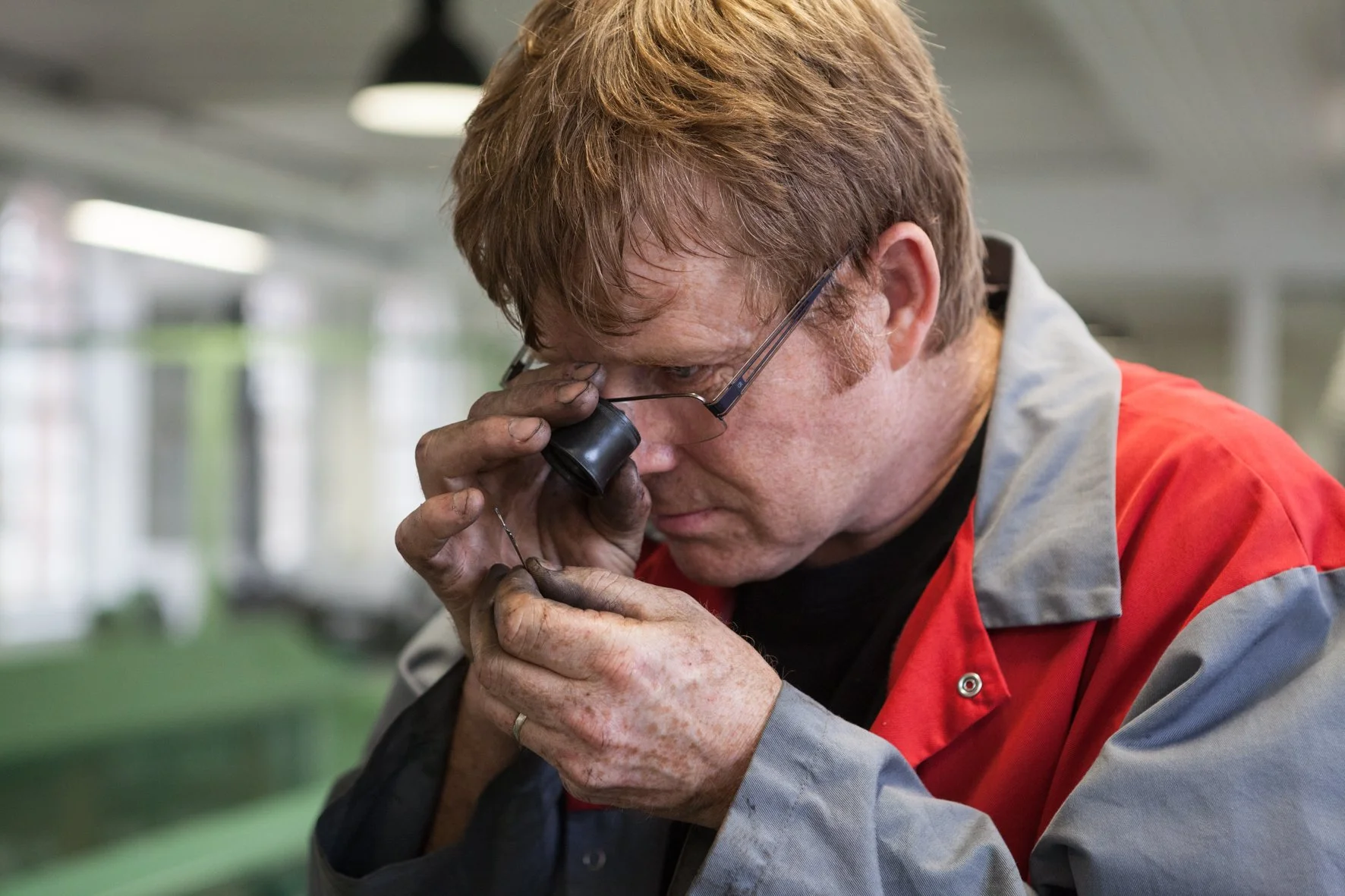 Un homme portant un blouson rouge et gris, examine de près quelque chose avec une loupe dans un environnement industriel ou d'atelier.