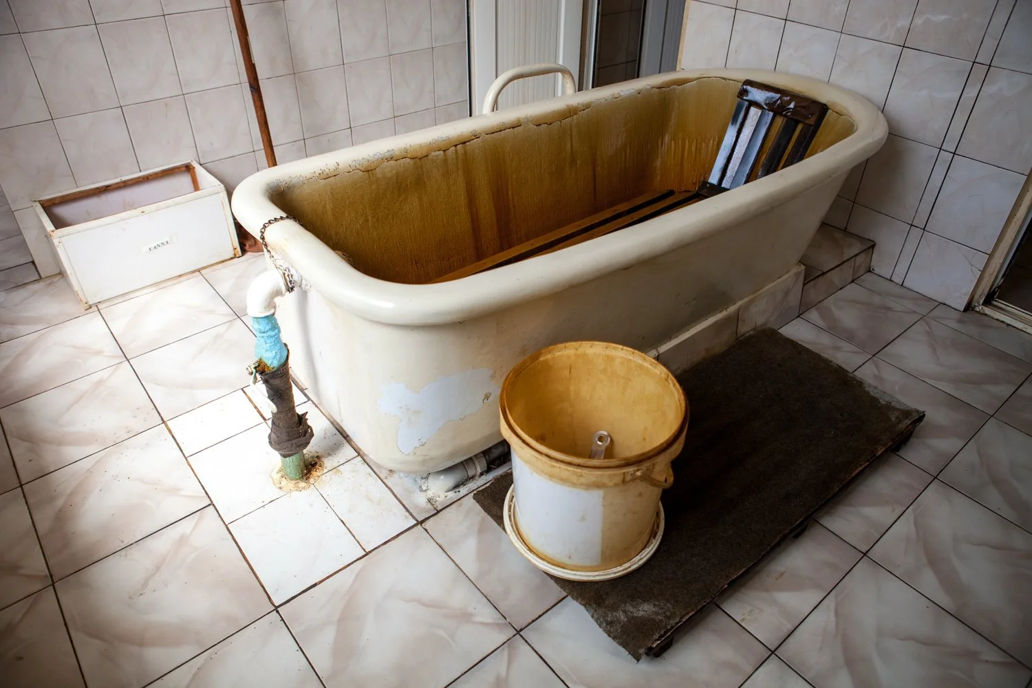 Une baignoire ancienne dans une salle de bain, avec un seau en plastique jaune à côté, des carreaux de sol en céramique blanche et un tapis noir.