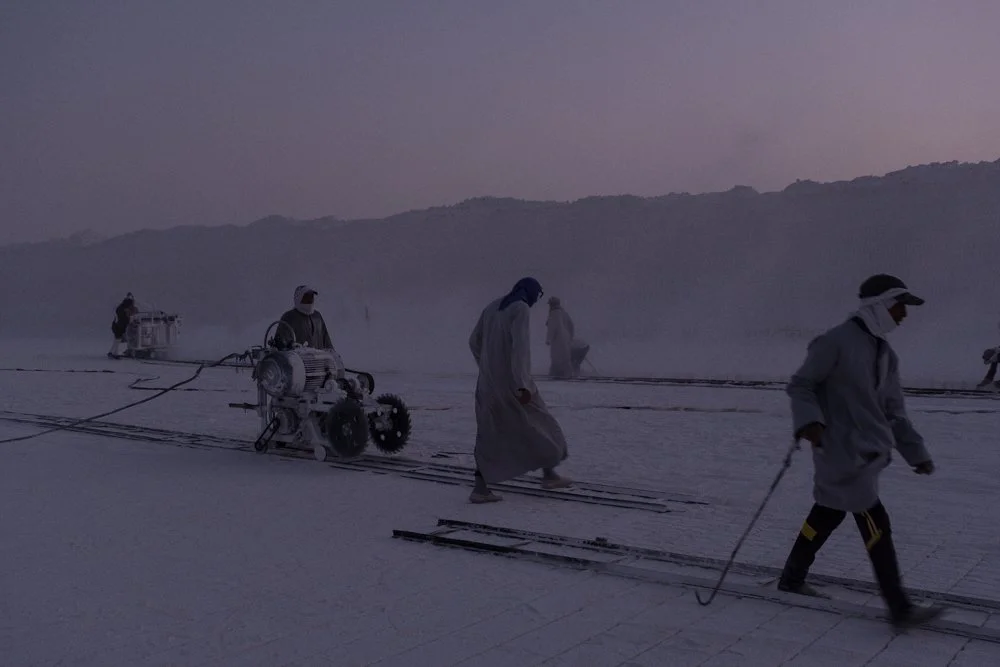 Des personnes travaillent dans une carrière de calcaire, en utilisant de grandes scies circulaires pour découper le sol, avec des montagnes en arrière-plan, au crépuscule.
