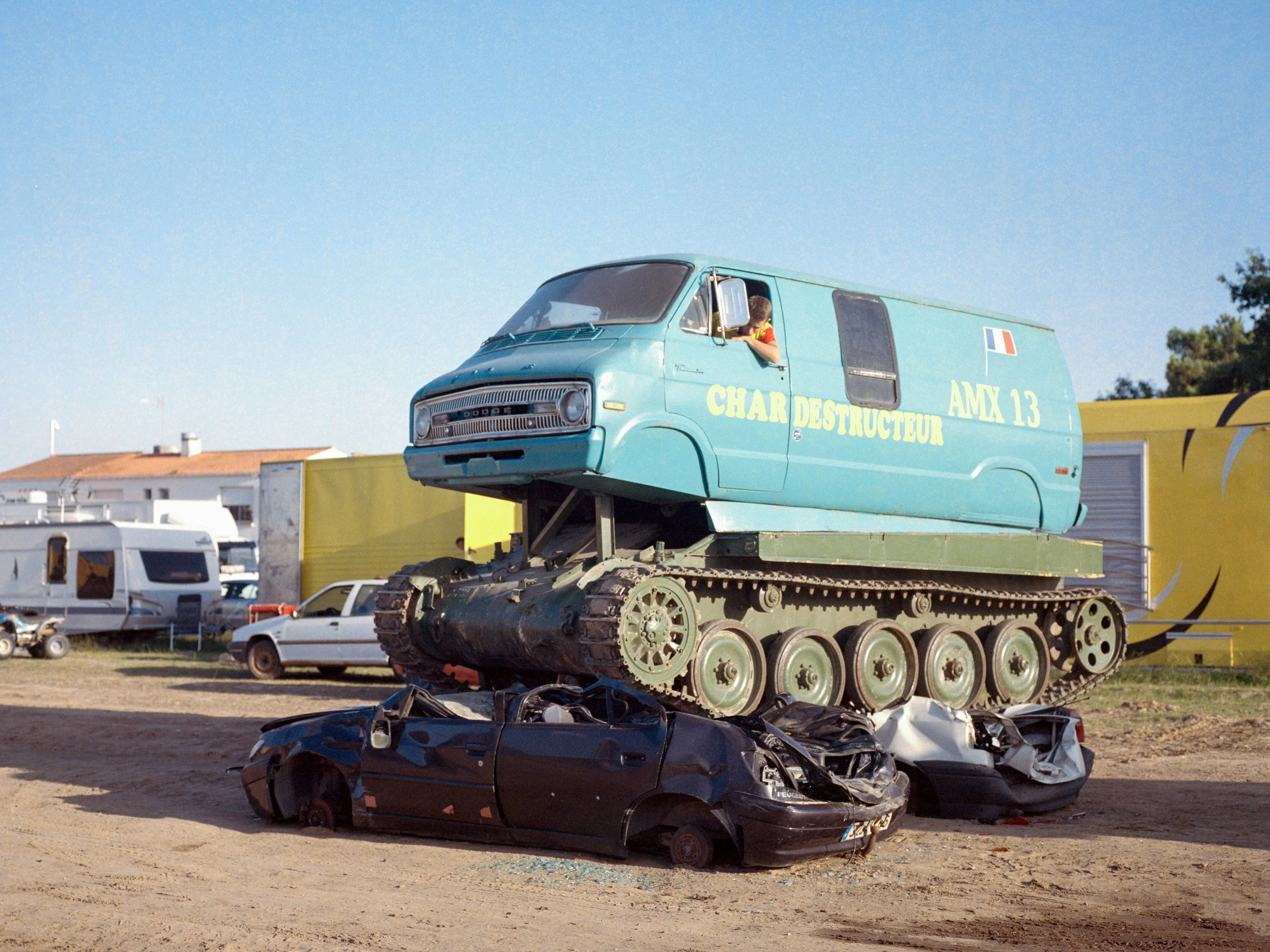 Un tank avec une carrosserie de van montant sur deux voitures écrasées, dans un environnement extérieur avec un ciel clair.