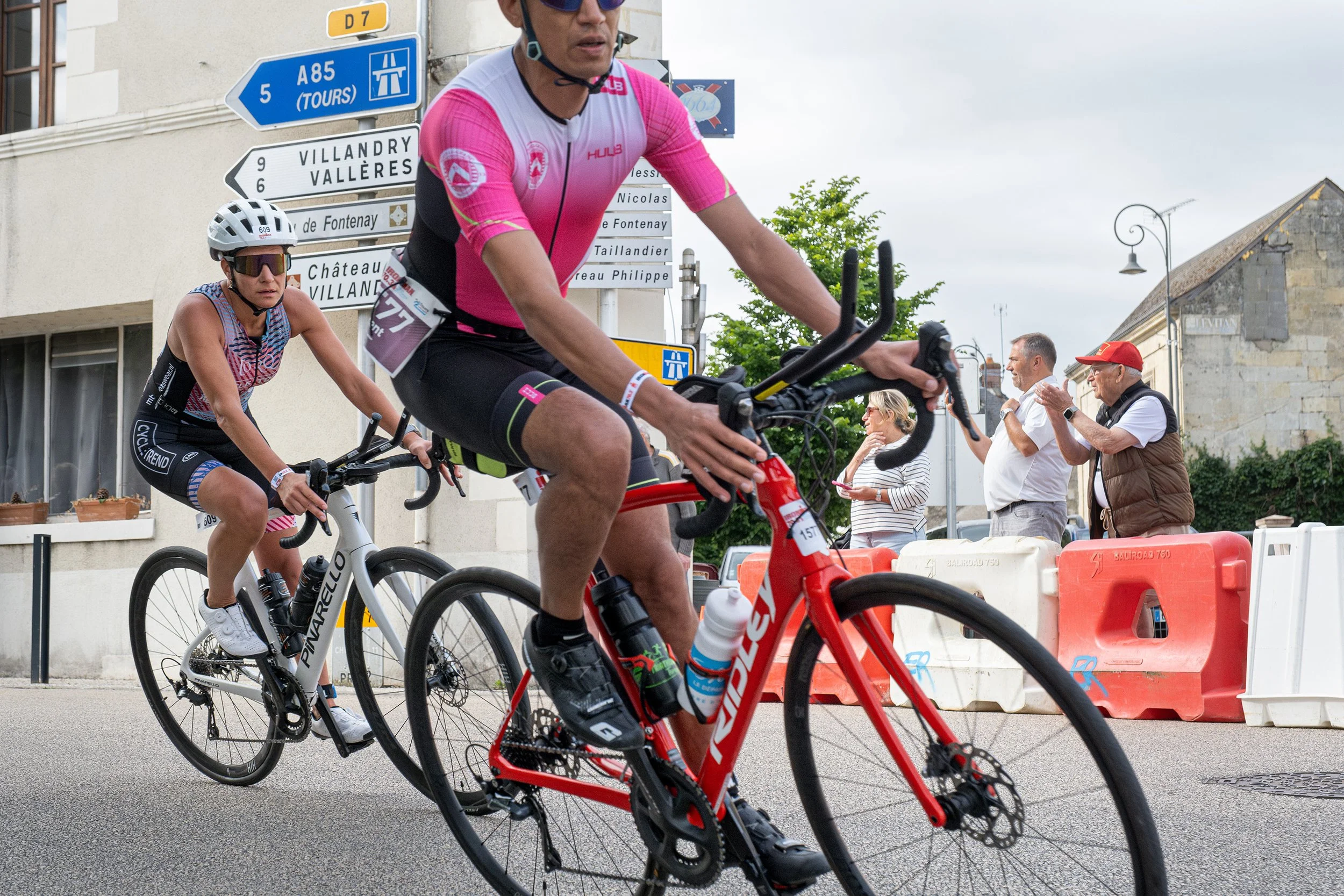 Deux cyclistes en compétition sur la route, avec des spectateurs applaudissant à côté. Un panneau de signalisation routière est visible en arrière-plan.