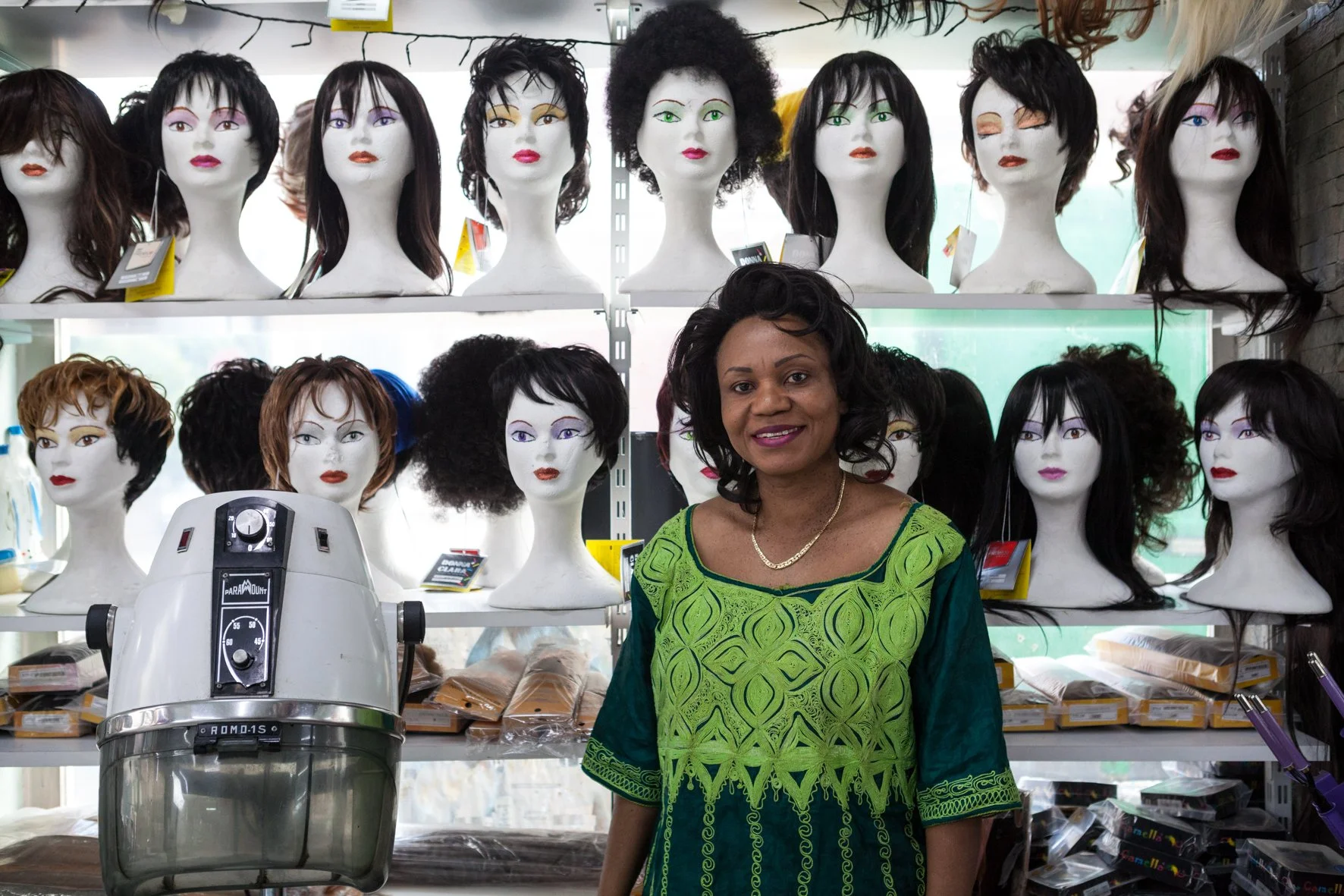 Une femme souriante se tient dans une boutique de perruques, entourée de plusieurs mannequins portant différentes perruques sur des étagères derrière elle.