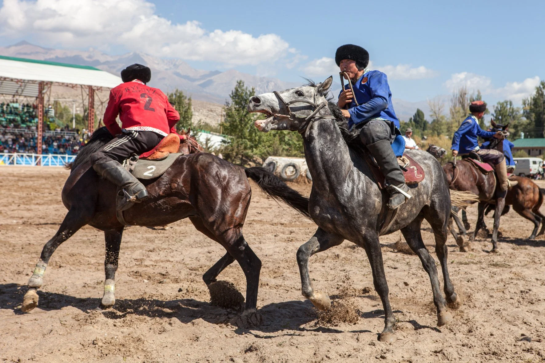 Participants en compétition de kok boru sur des chevaux dans un terrain en plein air, sous un ciel partiellement nuageux, avec des spectateurs en arrière-plan et des montagnes en toile de fond.