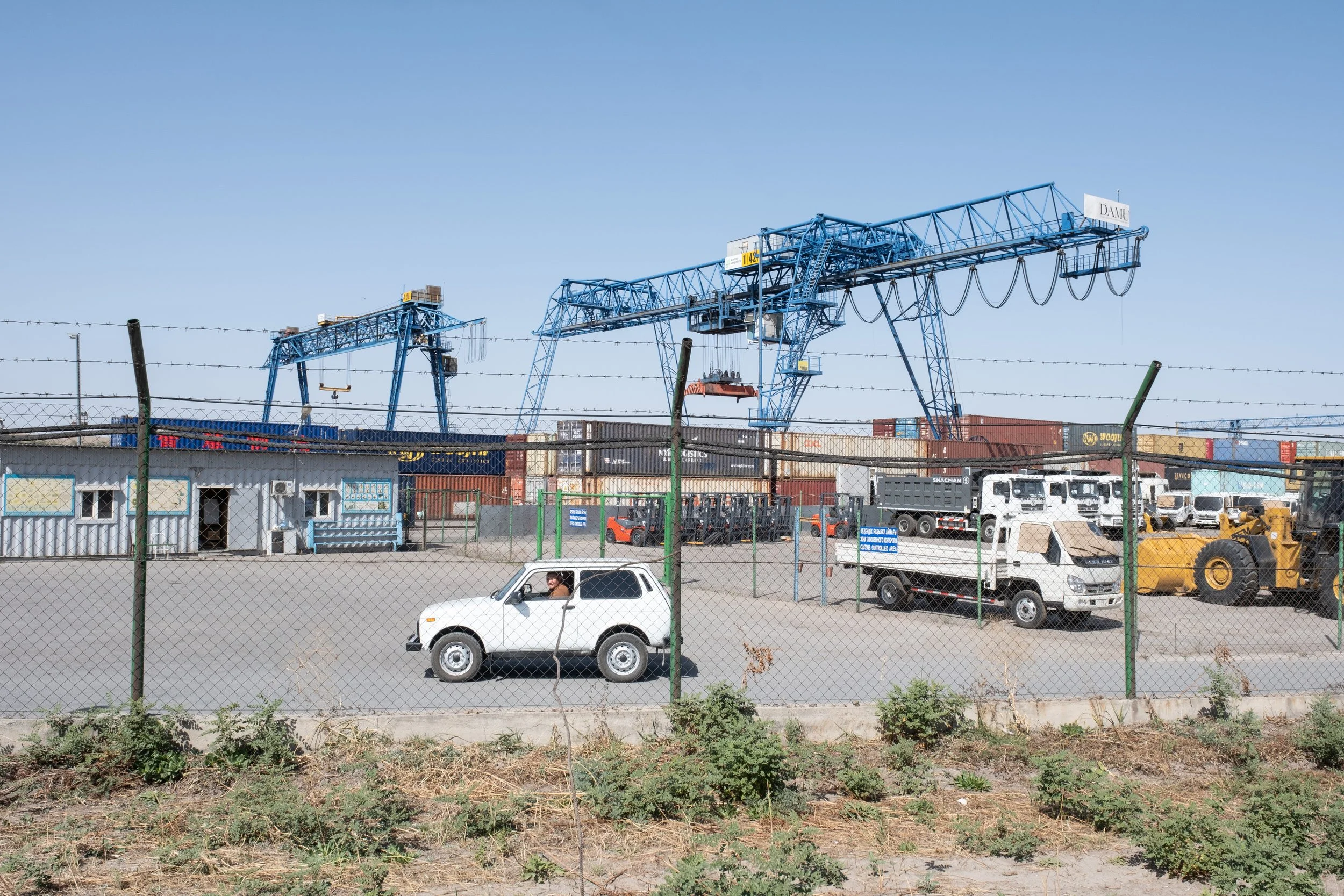 Zone portuaire avec plusieurs grues bleues, véhicules de chantier et containers, entourée d'une clôture en fer et de quelques bâtiments, sous un ciel clair.