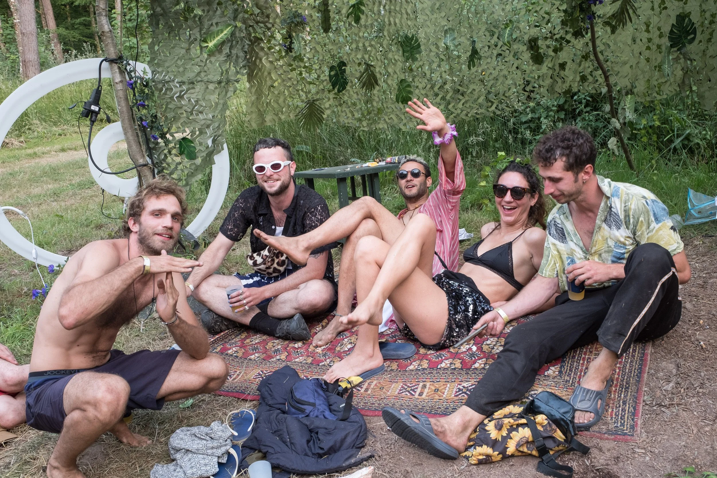 Groupe de six amis en maillot de bain et vêtements décontractés assis et allongés sur un tapis dans un espace extérieur verdoyant, souriants et détendus.