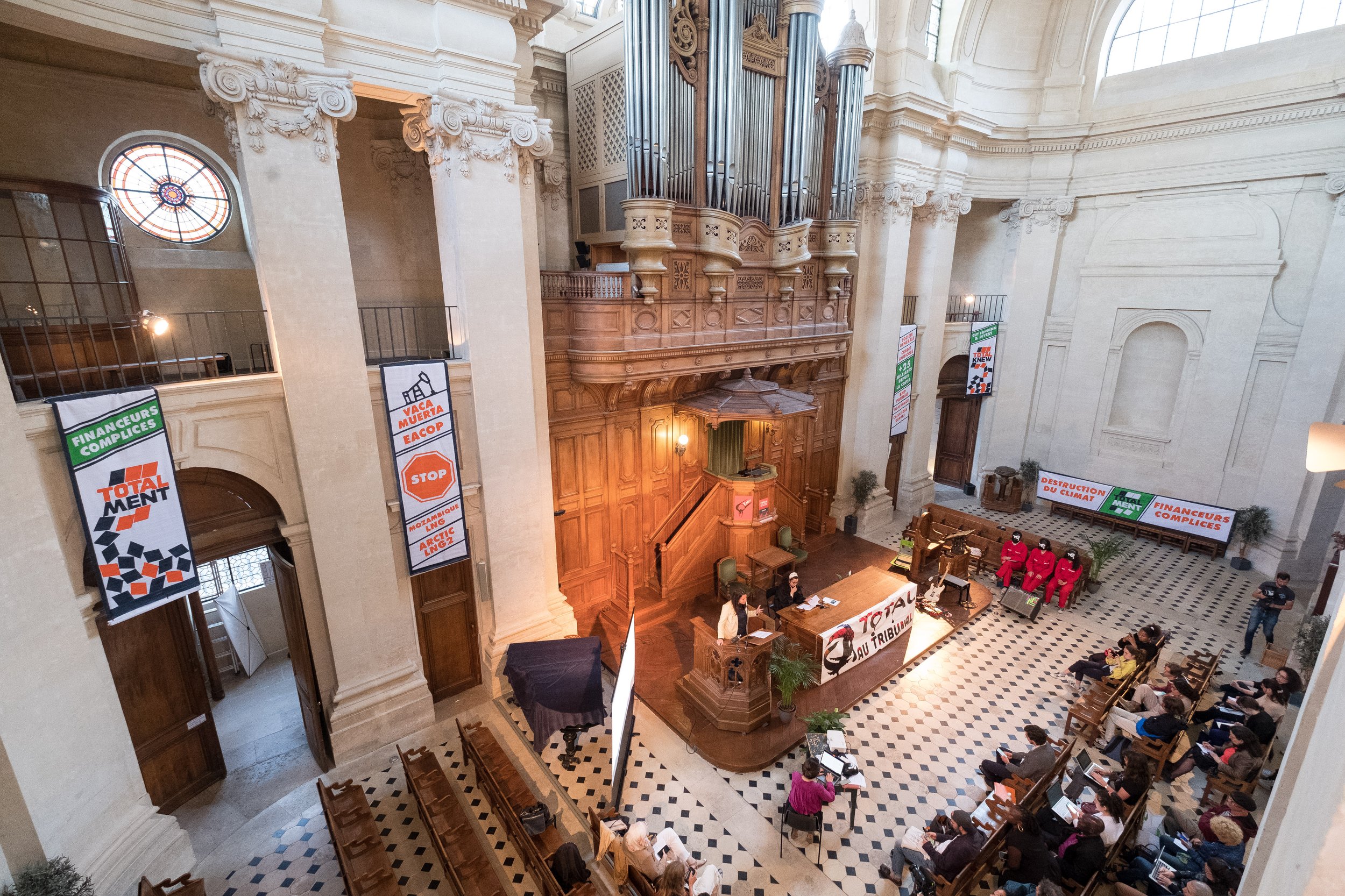 Une salle d'audience avec des gradins occupés par des personnes, un podium avec une banderole « TOTAL AU TRIBUNAL », et des pancartes avec des slogans comme « Destruction du climat » et « Financiers complices ».