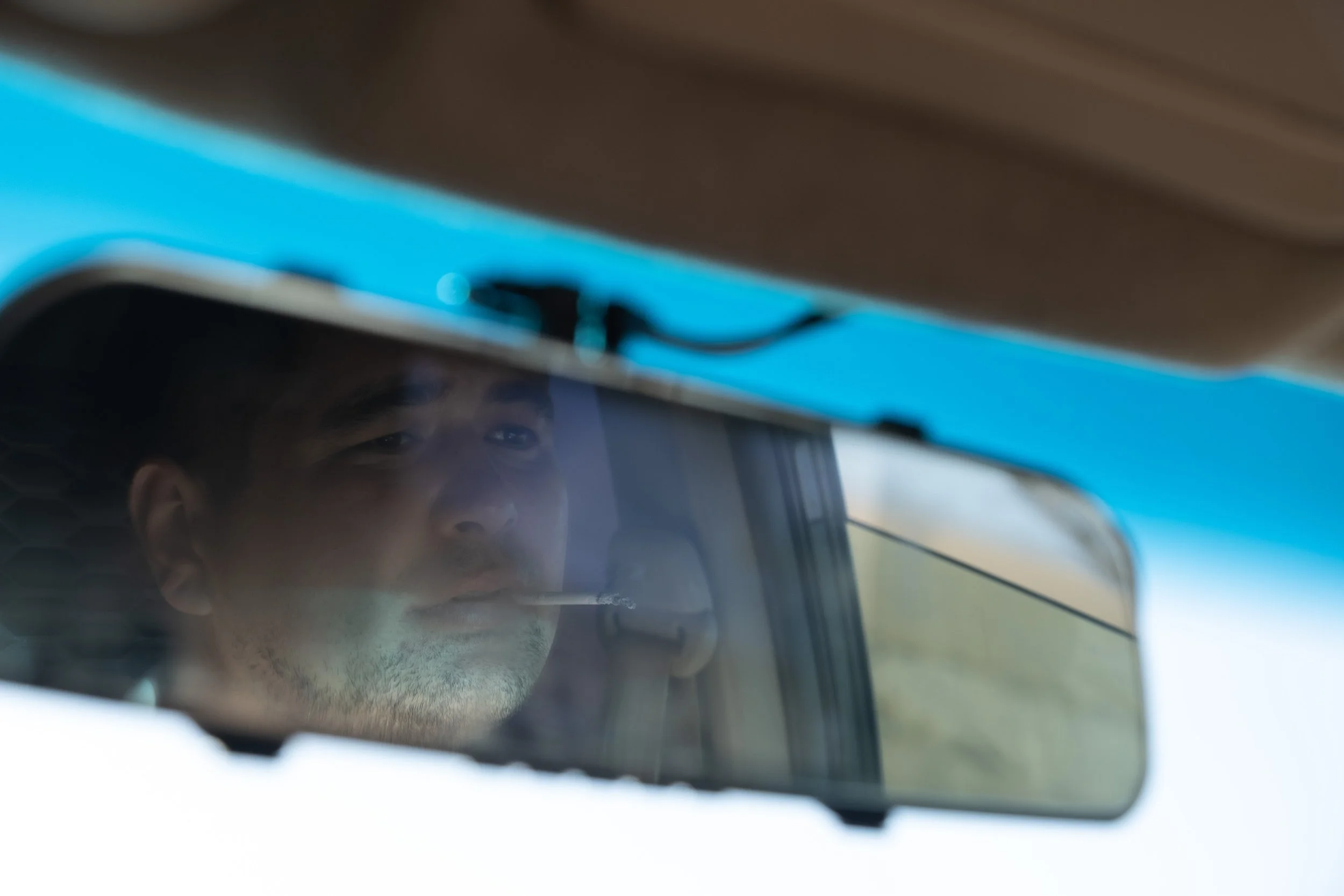 Photo d'un homme à travers le rétroviseur de la voiture, avec un cigarette à la bouche.