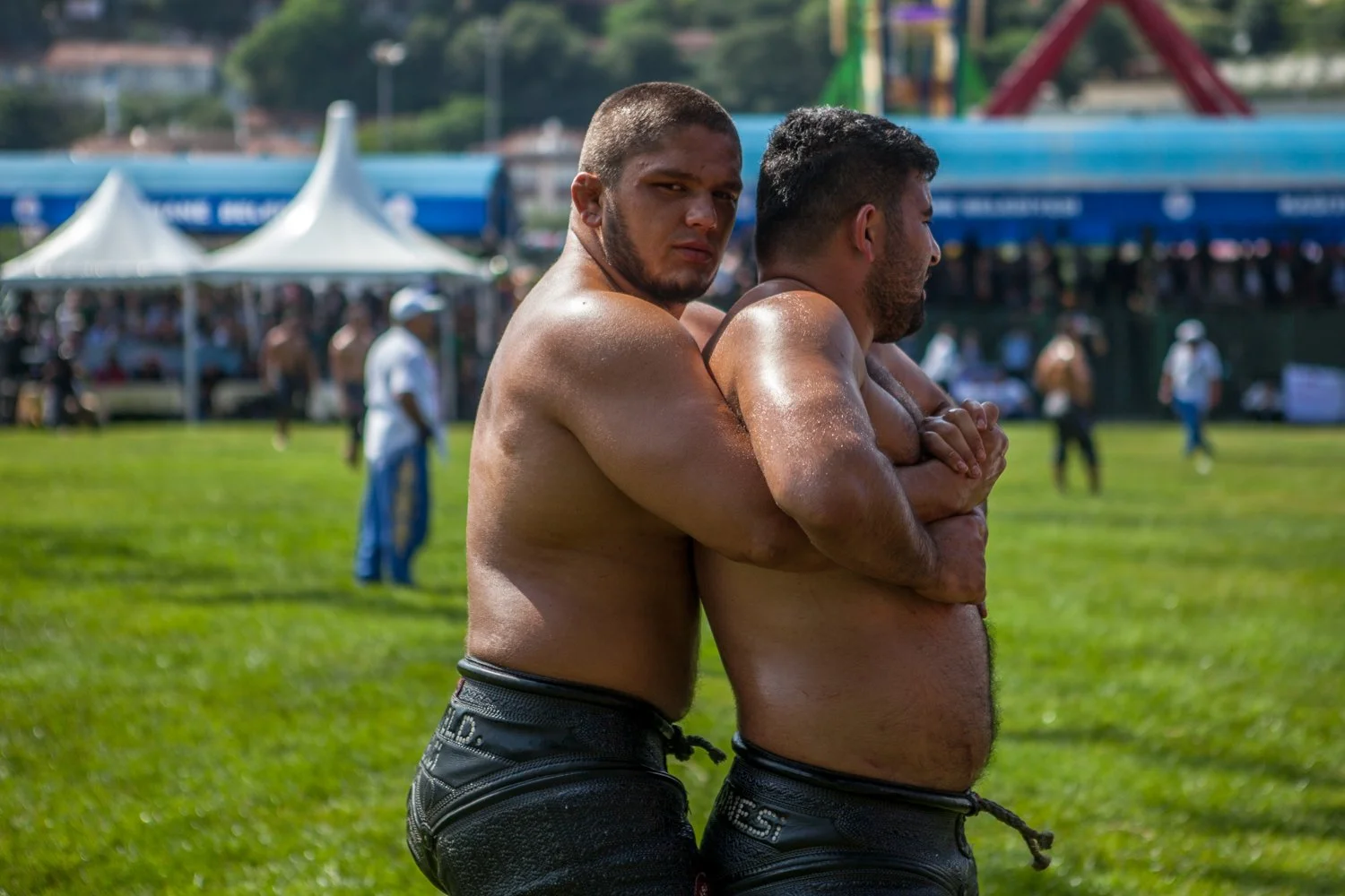 Deux hommes nus, trempés de sueur, en position de lutte sur une grande pelouse verte, avec un public et des structures de fête en arrière-plan.