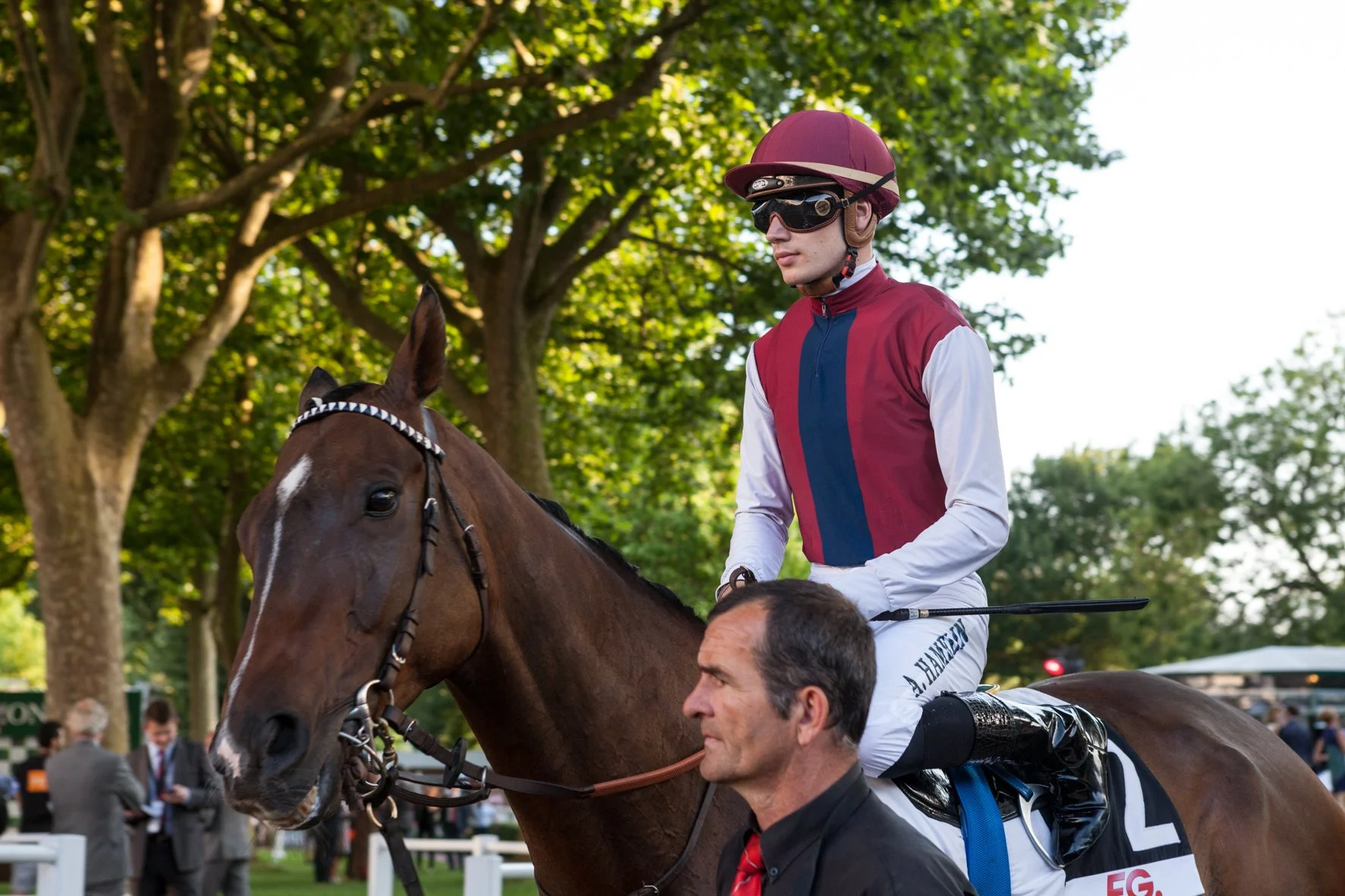 Jockey en tenue de course avec casque et lunettes, assis sur un cheval avant une course, avec un homme en costume en arrière-plan et des arbres en fond.