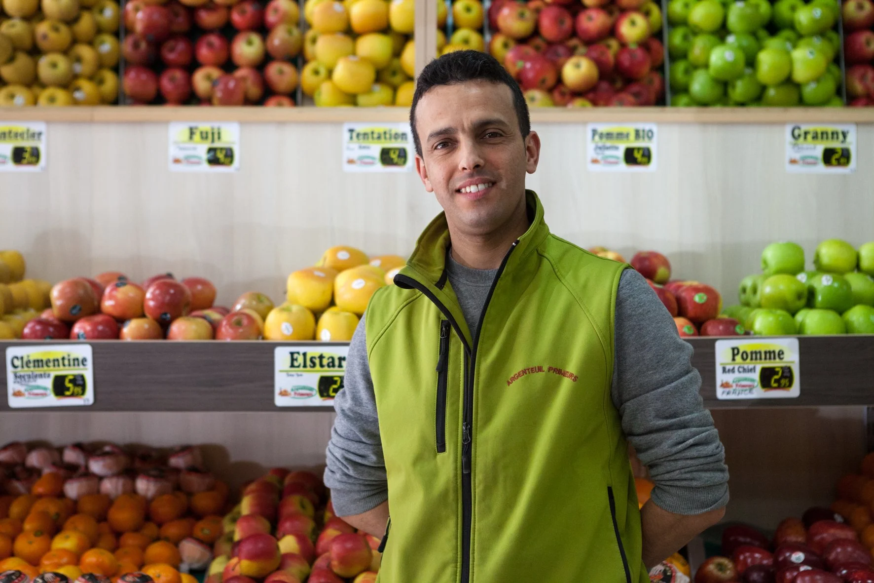 Un homme en veste verte se trouve dans une épicerie, avec des étagères de fruits derrière lui, notamment des pommes et des oranges.