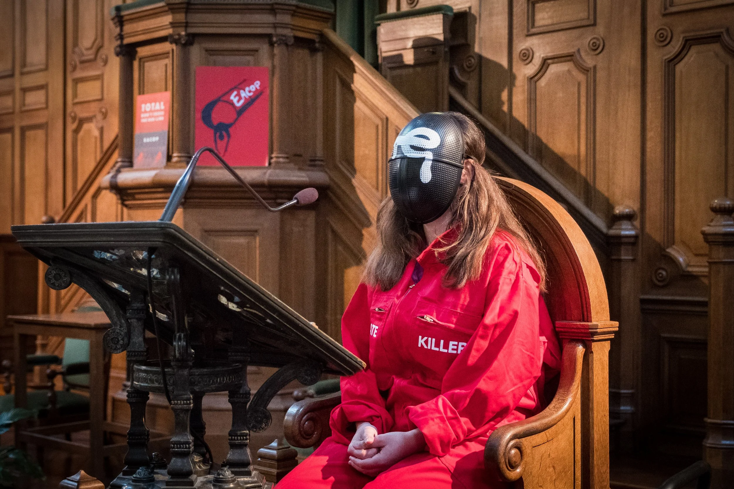 Une femme militante de 350.org portant un masque noir  et une veste rouge avec des mots en anglais, assise dans une salle en bois, derrière un pupitre.
