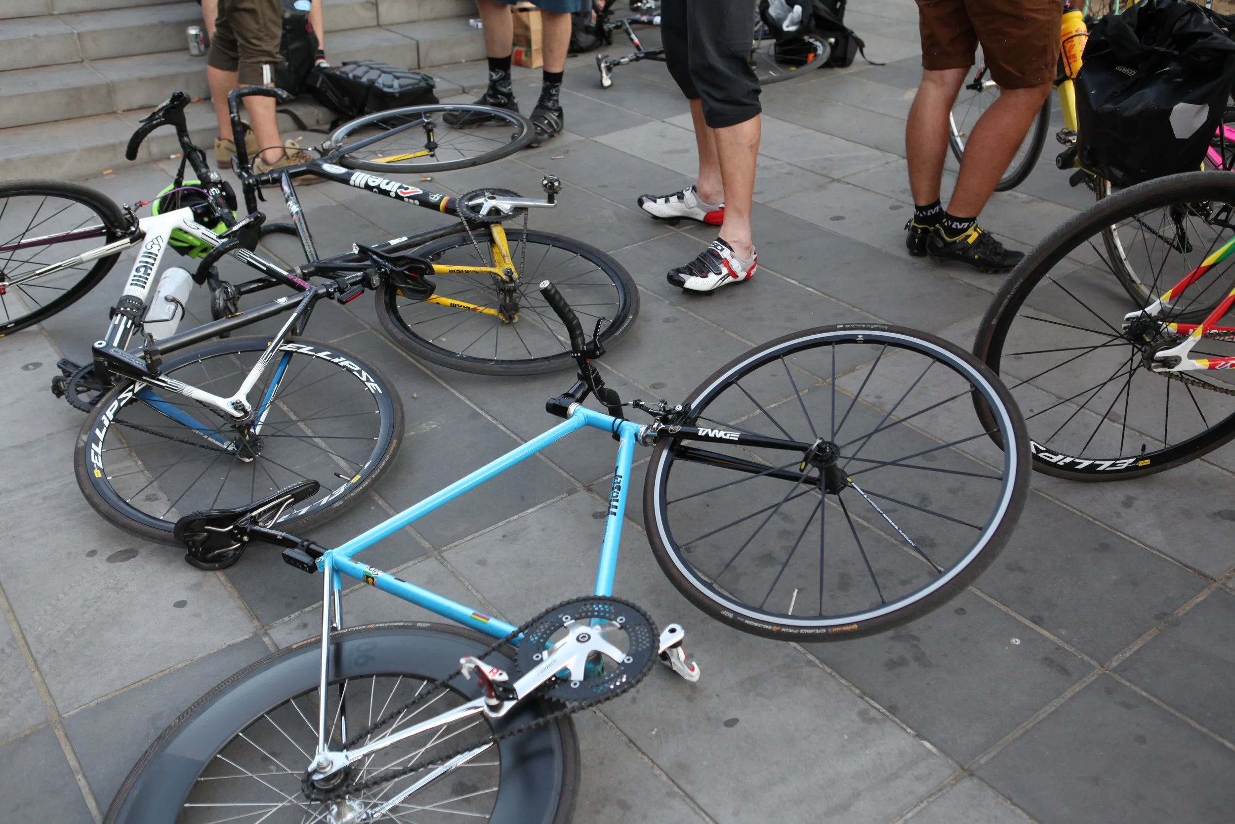Plusieurs vélos allongés ou stationnés sur un sol pavé, avec des personnes debout autour, portant des chaussures de sport.
