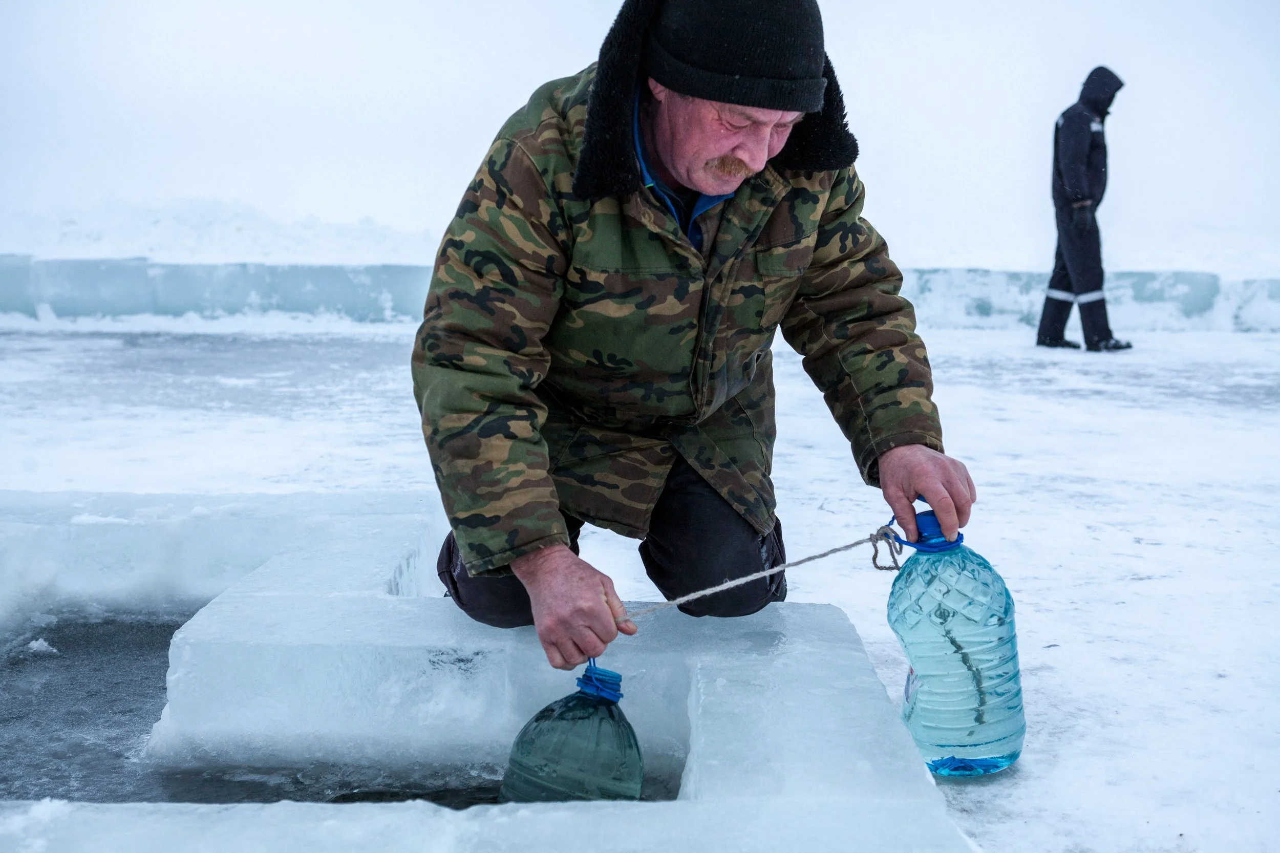 Un homme en habits militaires en train de remplir deux bouteilles d'eau à partir d'un trou dans la glace, dans un environnement glacé avec de la neige, en Russie.