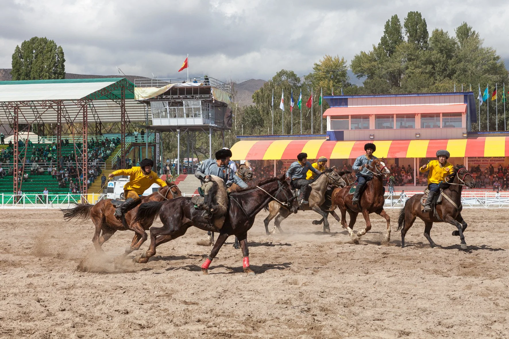 Une course de cheval traditionnel en Asie centrale avec des cavaliers, sur une piste en terre à l'extérieur. L'arrière-plan montre des gradins avec des spectateurs, des drapeaux multicolores dans un ciel nuageux.