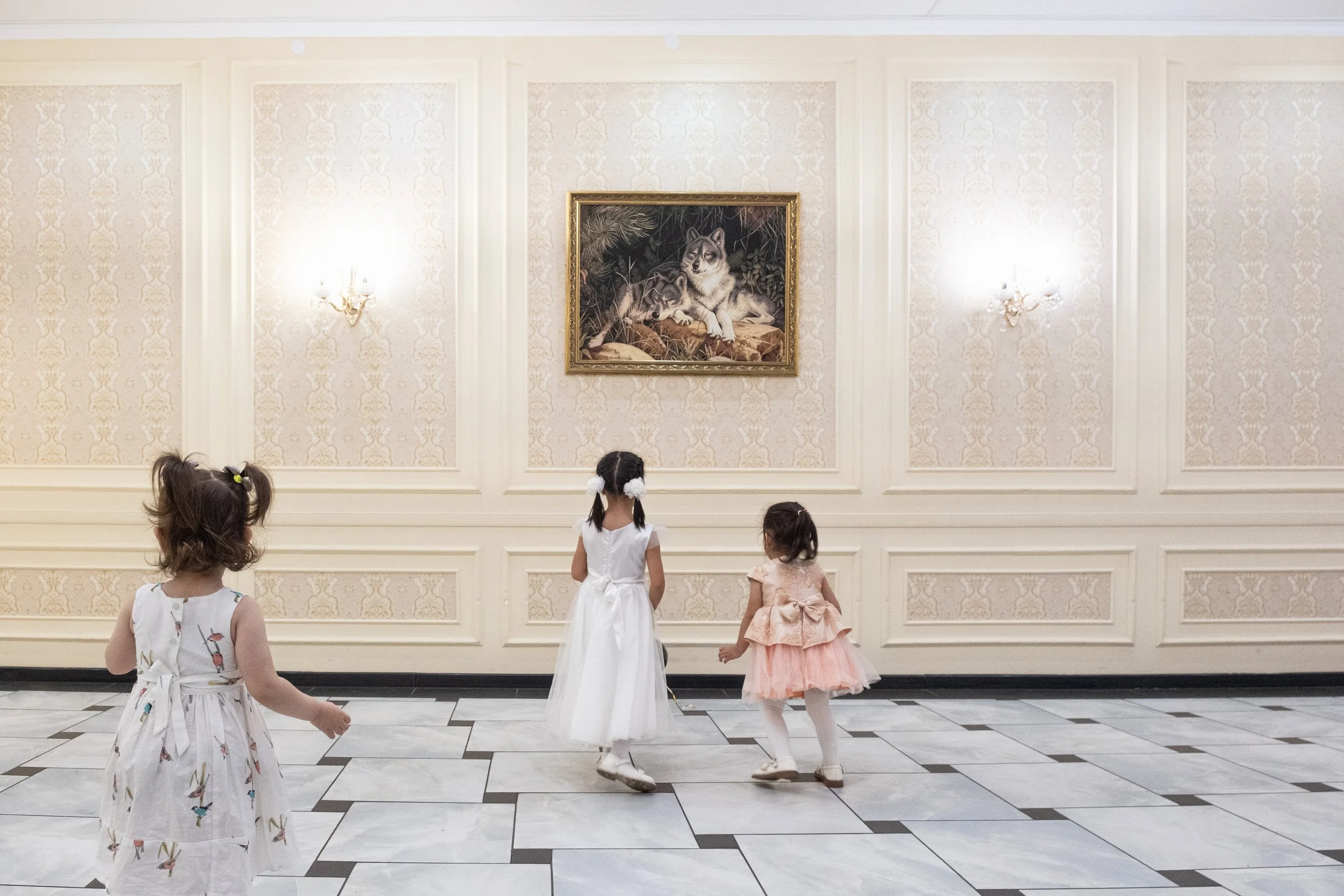Trois jeunes filles en robes blanches et roses regardant un tableau de loup dans une salle des fêtes élégante au Kazakhstan.