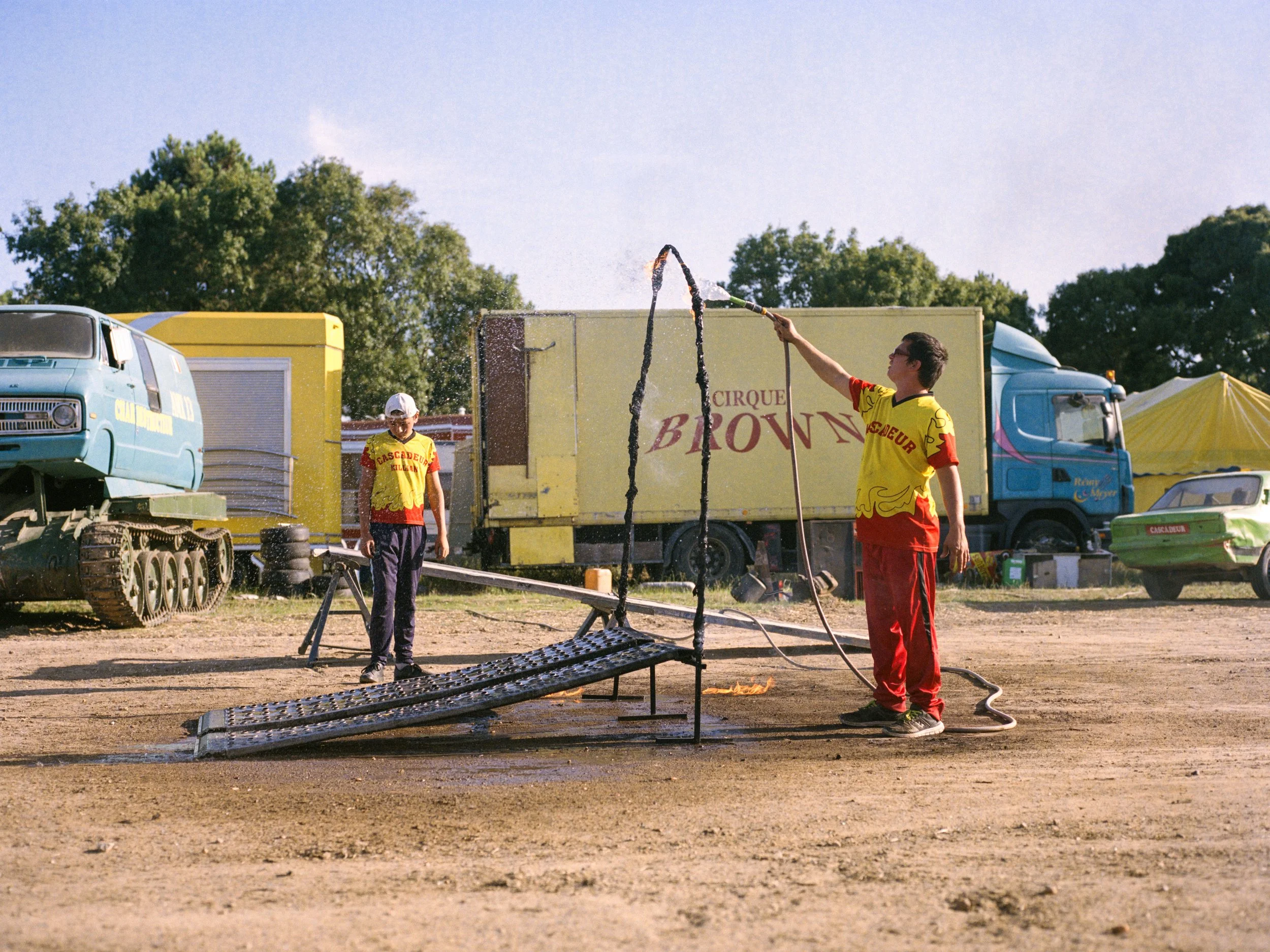 Deux hommes en vêtements de cirque arrosent un arceau métallique avec un tuyau d'arrosage. Un camion jaune avec l'inscription « Cirque Brown » en arrière-plan, entouré de tentes colorées et d'autres véhicules.
