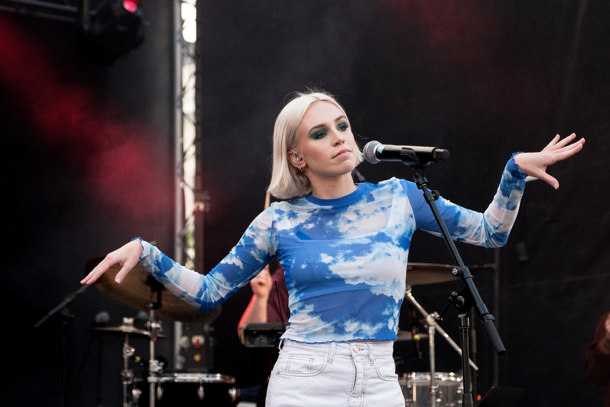 Une femme blonde avec des yeux maquillés en bleu chante sur scène avec un micro, portant un haut à motif ciel avec des nuages et un pantalon blanc, lors d'une performance en direct.