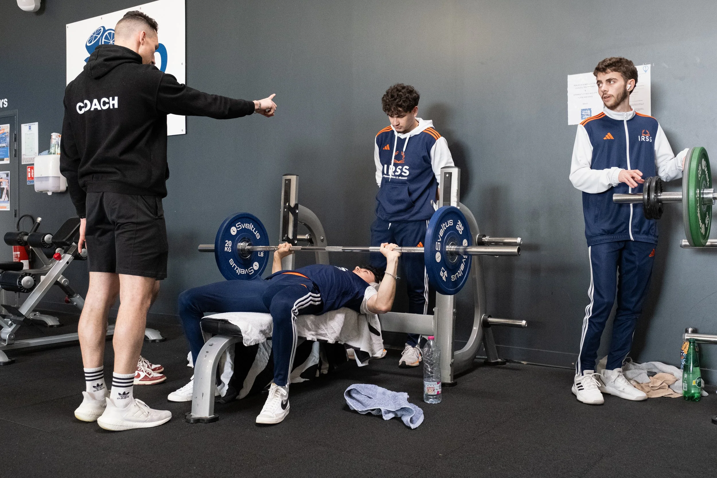 Un entraîneur de coach donne des instructions à deux jeunes hommes qui font un développé couché dans une salle de sport. Un jeune homme est allongé sur un banc soulevant une barre avec des poids, tandis que l'autre se tient à côté, prêt à l'aider. Un