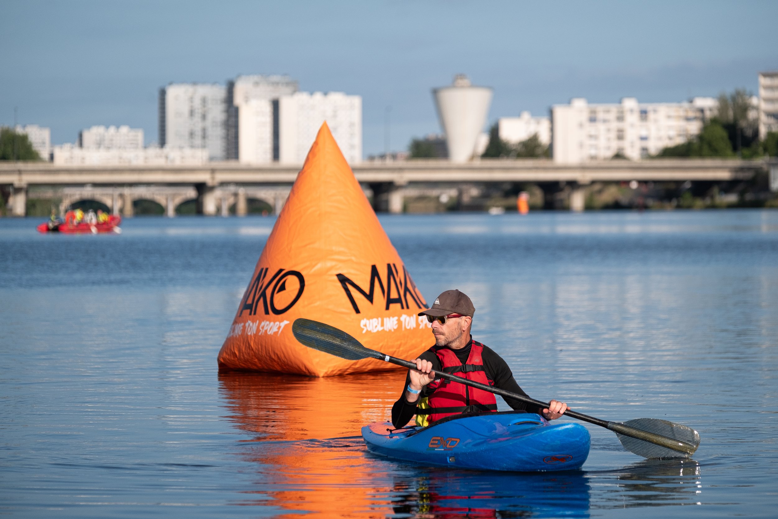 Un homme en kayak sur une rivière, portant un gilet rouge, avec une balise orange flottante derrière lui. En arrière-plan, un pont et des bâtiments urbains.