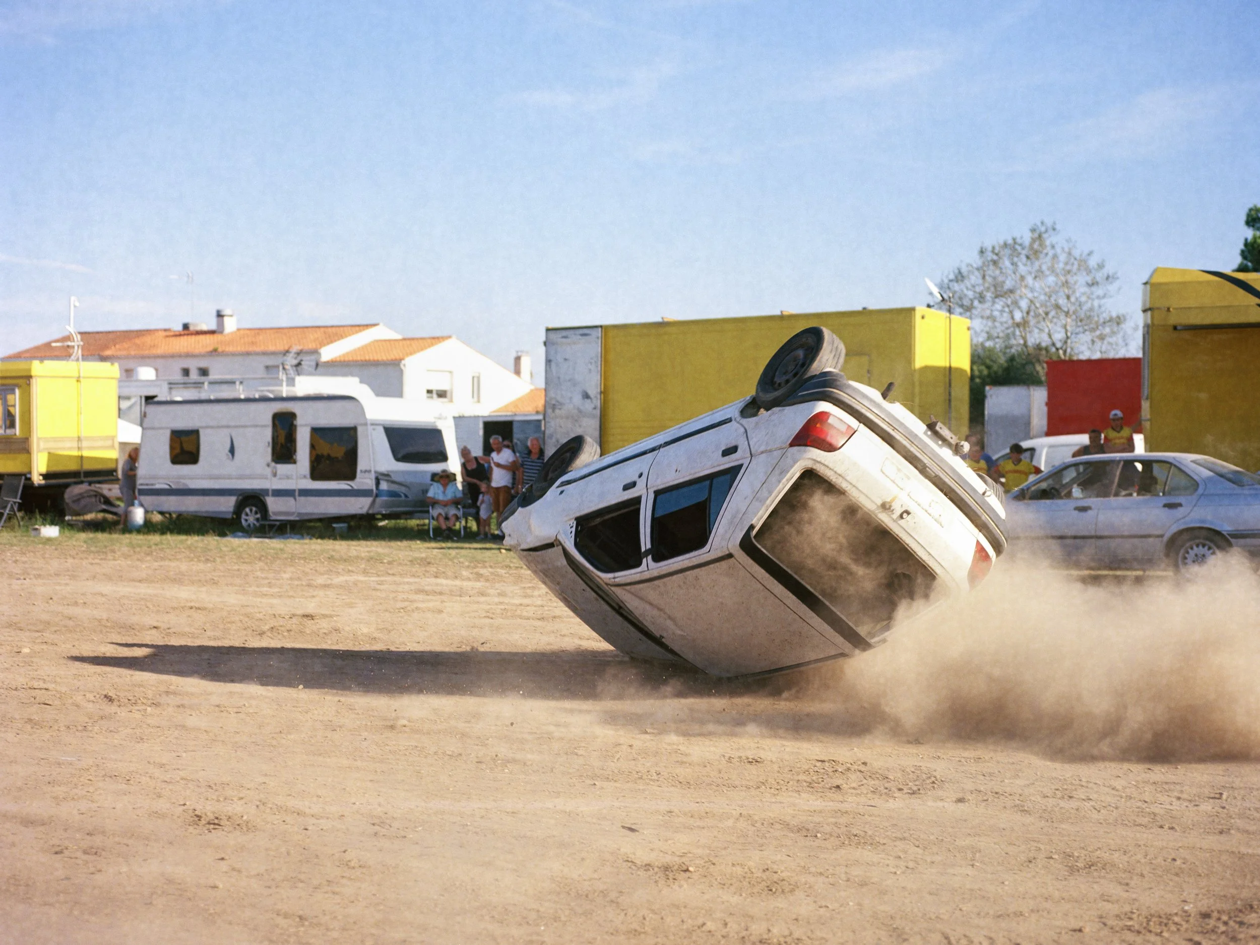 Une voiture blanche en train de faire un tonneau sur un terrain poussiéreux, avec des spectateurs et des camions en arrière-plan.